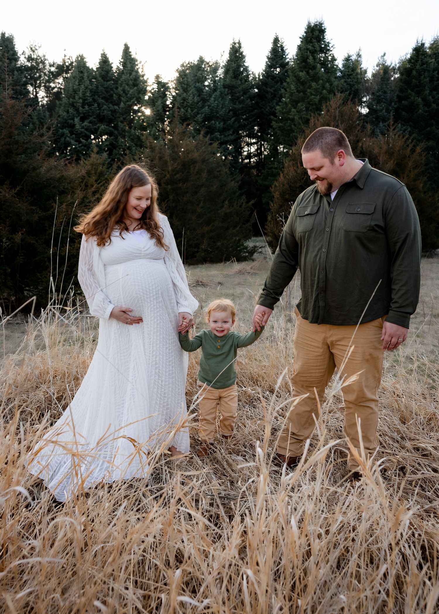 Pregnant woman in white dress holds toddler's hand with husband in autumn field.