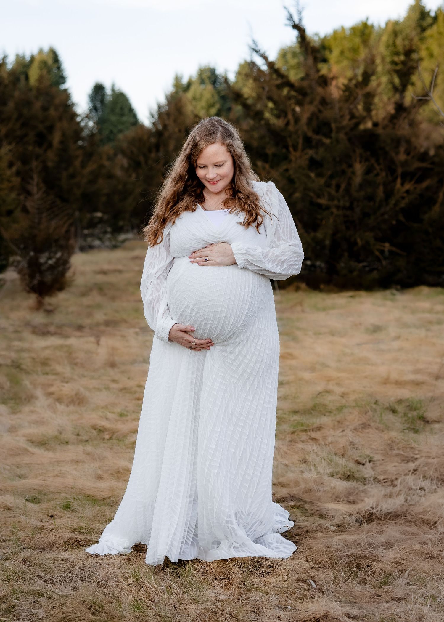 Glowing pregnant woman in flowing white lace gown smiling down at baby bump outdoors.