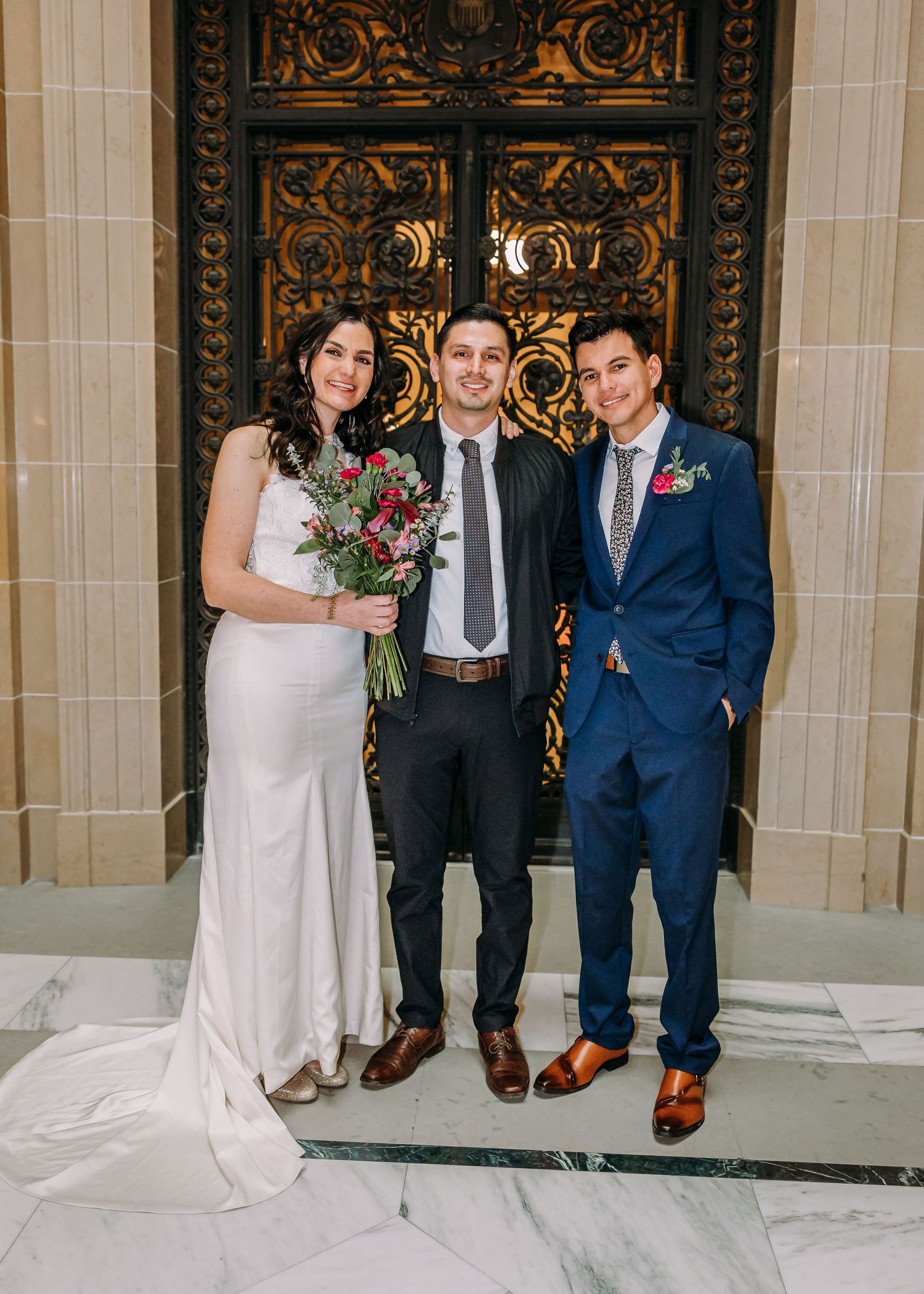 A bride holding flowers poses with two men in suits before golden ornate doors at a wedding.