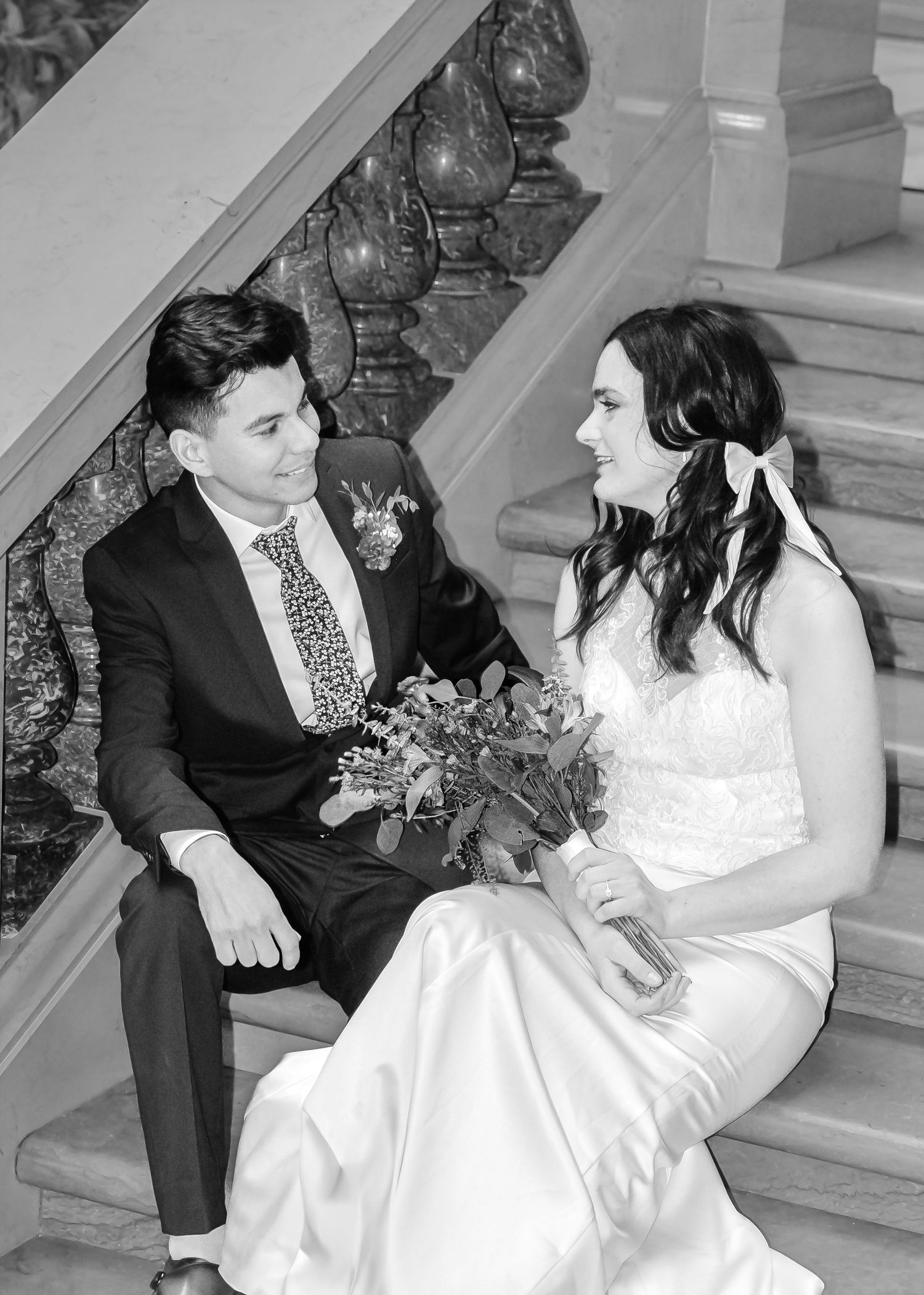 Black and white photo of bride and groom sitting on grand staircase steps, smiling at each other on wedding day.