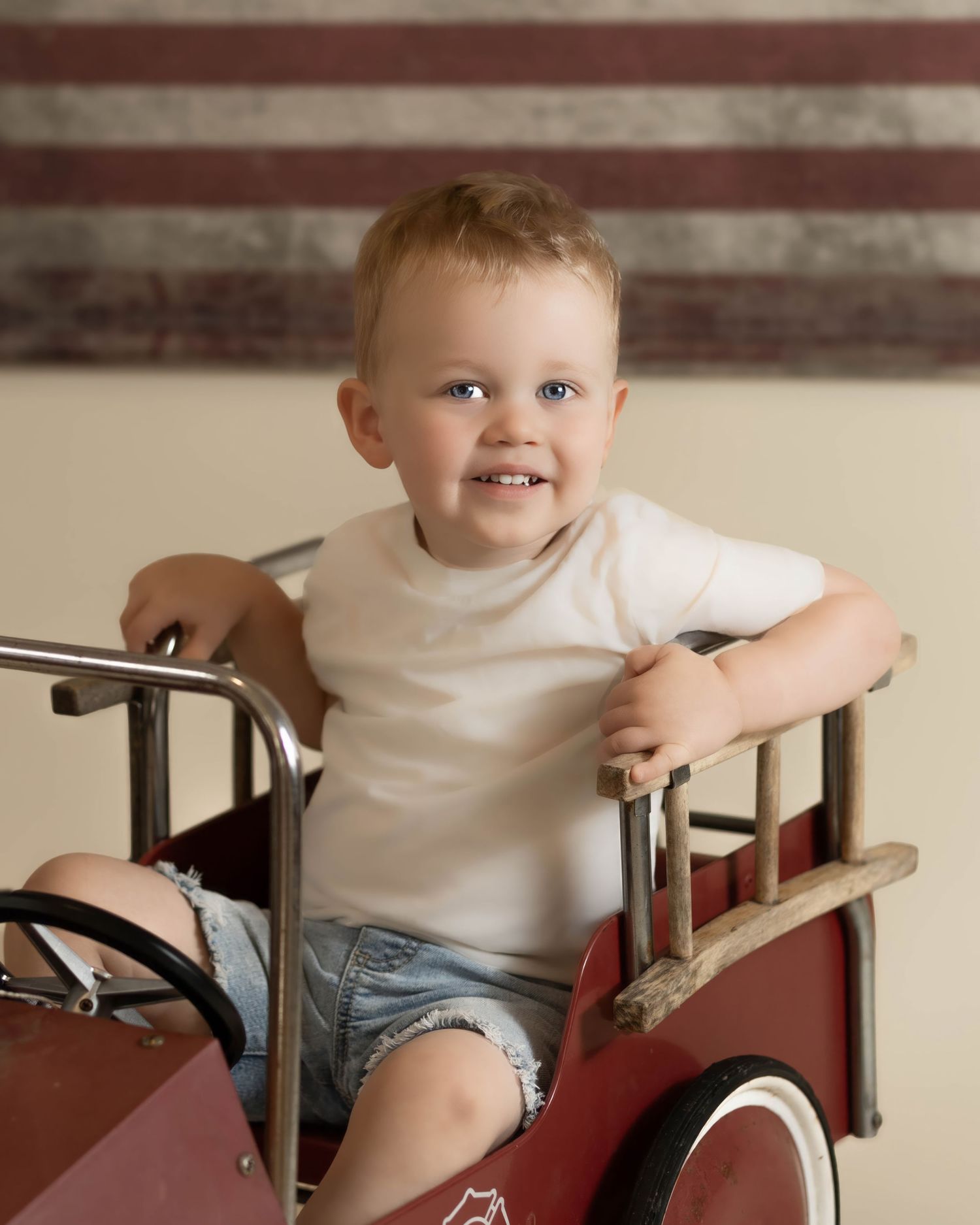 Smiling toddler in white shirt sitting in a vintage red pedal fire truck.