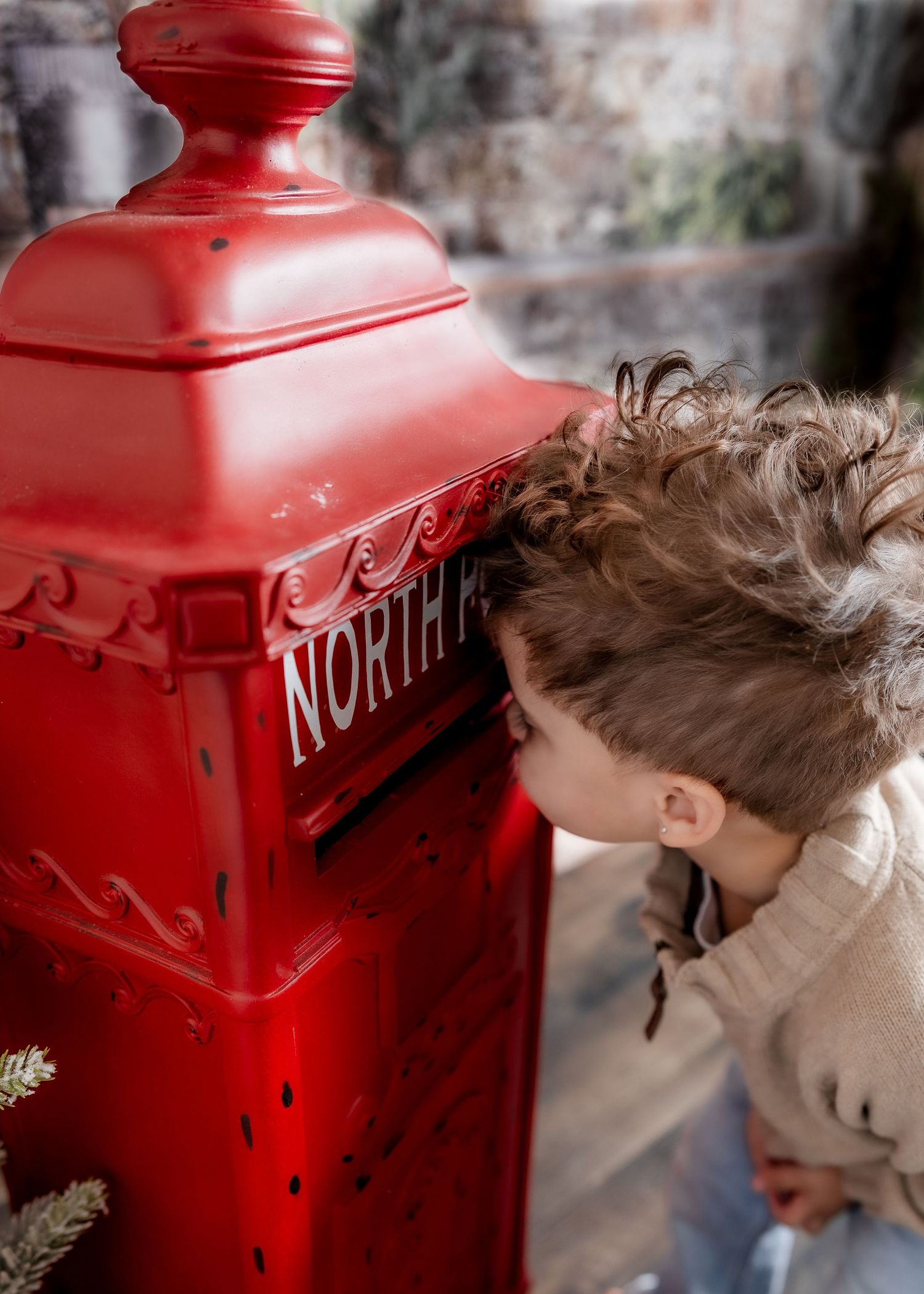 A curly-haired child in a beige sweater leans in to peek inside a red North Pole Christmas mailbox.