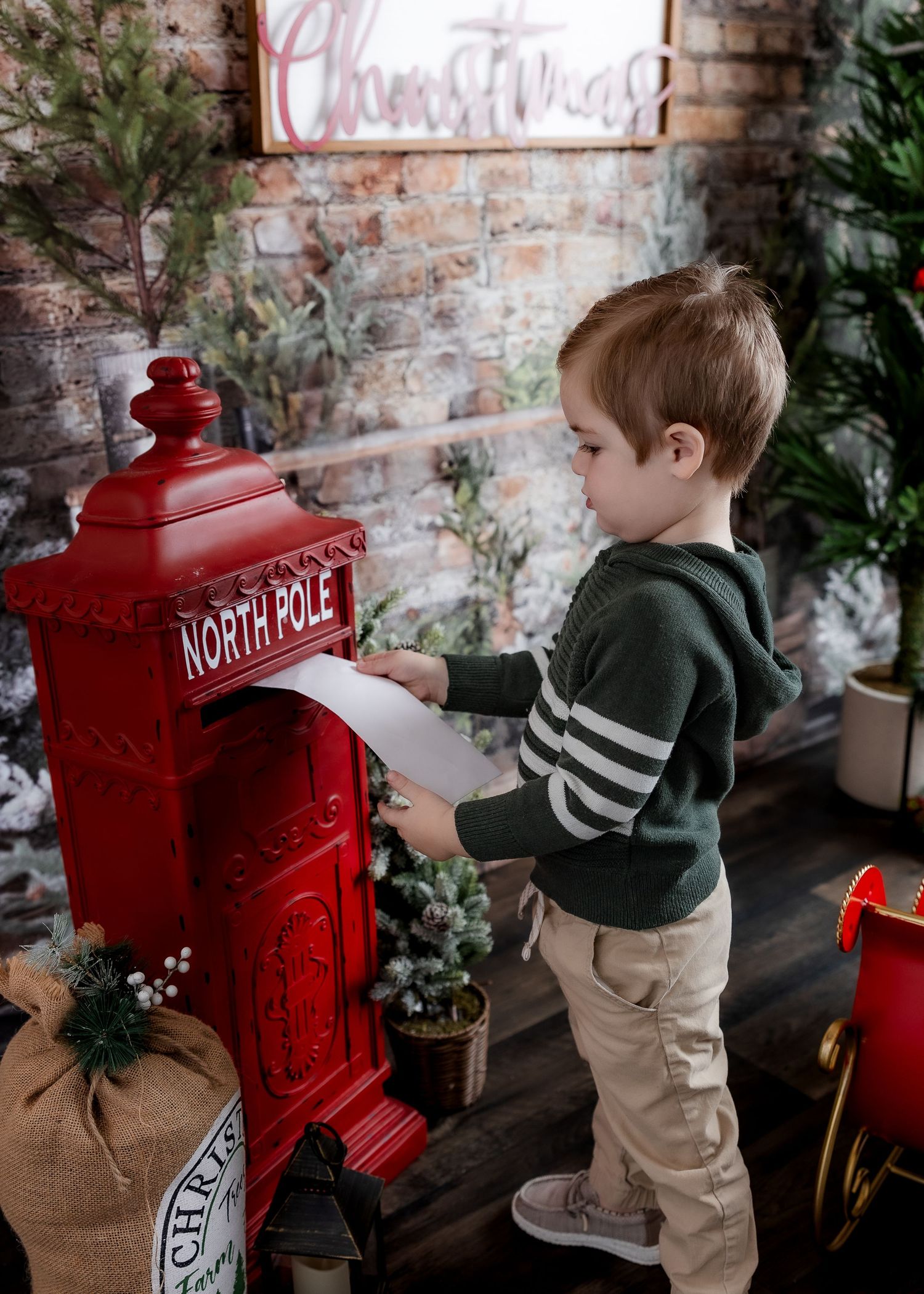 A young boy in a green sweater mailing a letter to Santa at a red North Pole mailbox in a Christmas setting.