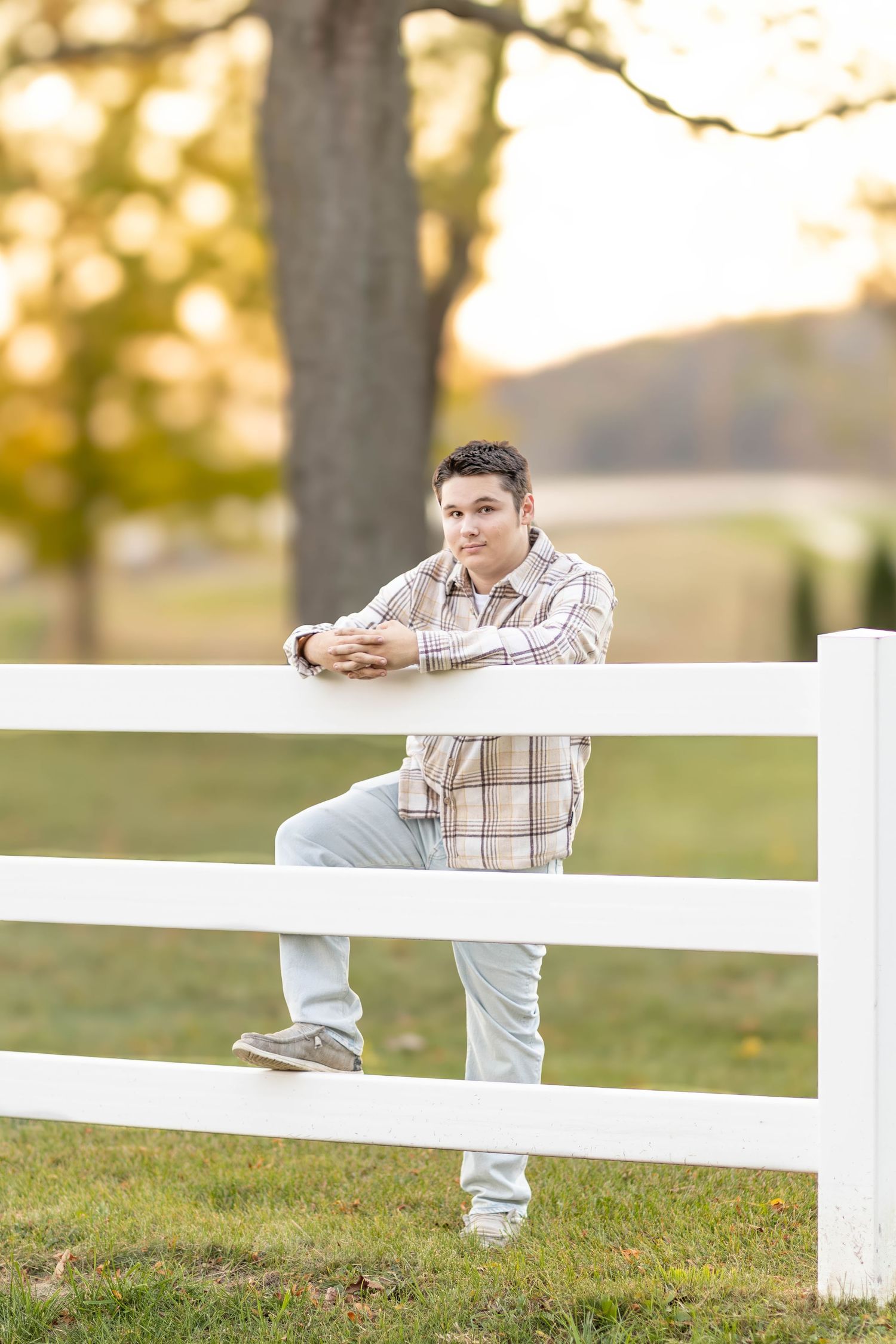 Senior boy in plaid shirt leans on white fence rail at sunset farm setting.