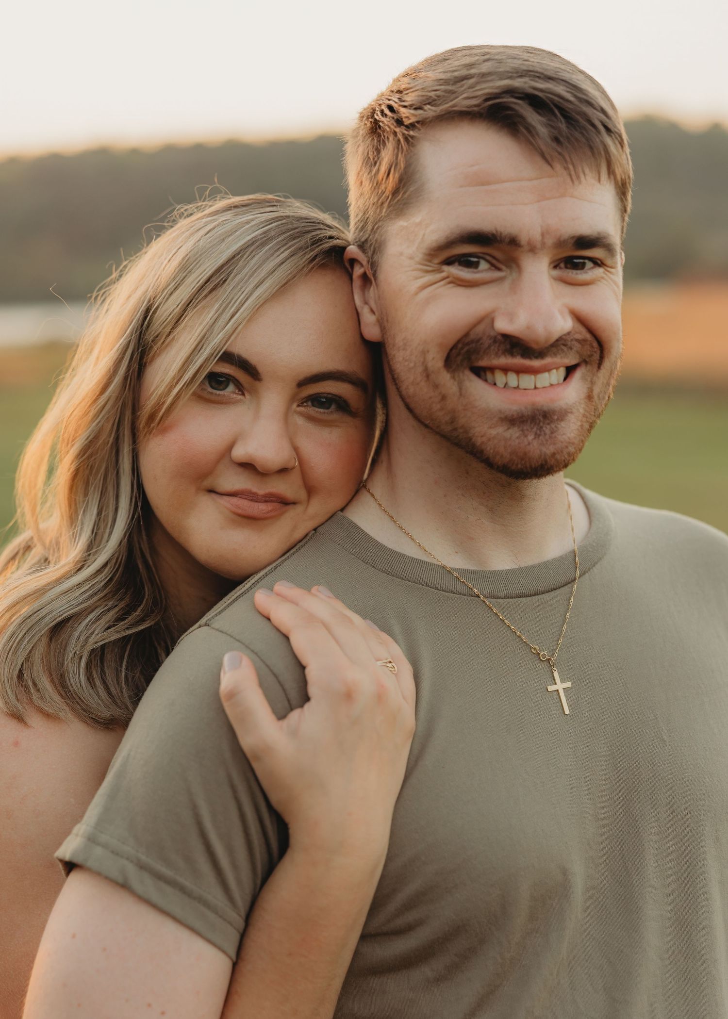 Smiling couple posing together at sunset, woman resting head on mans shoulder outdoors.