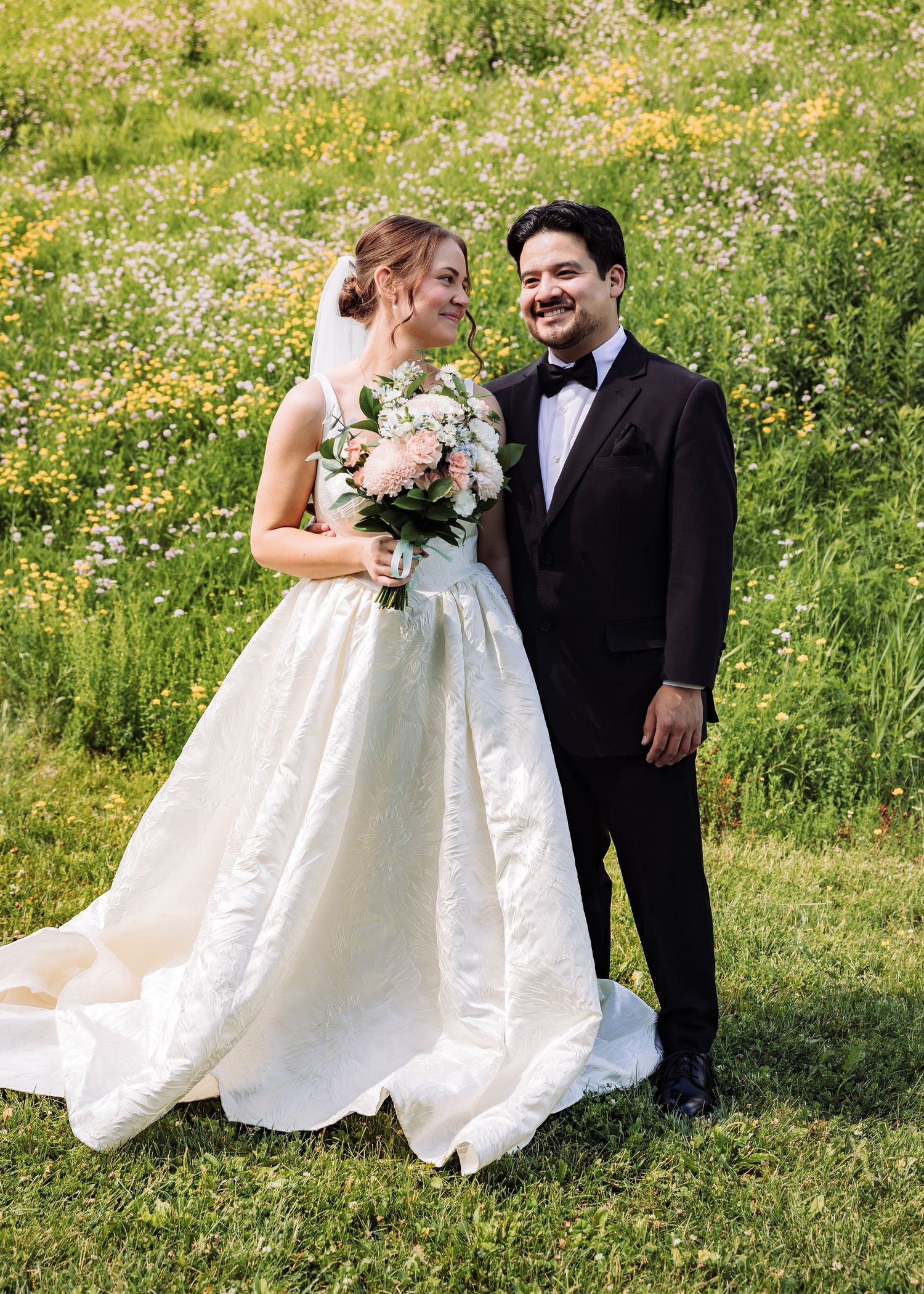 A bride in a white ball gown holding a floral bouquet stands with a groom in a black tuxedo in a wildflower meadow.