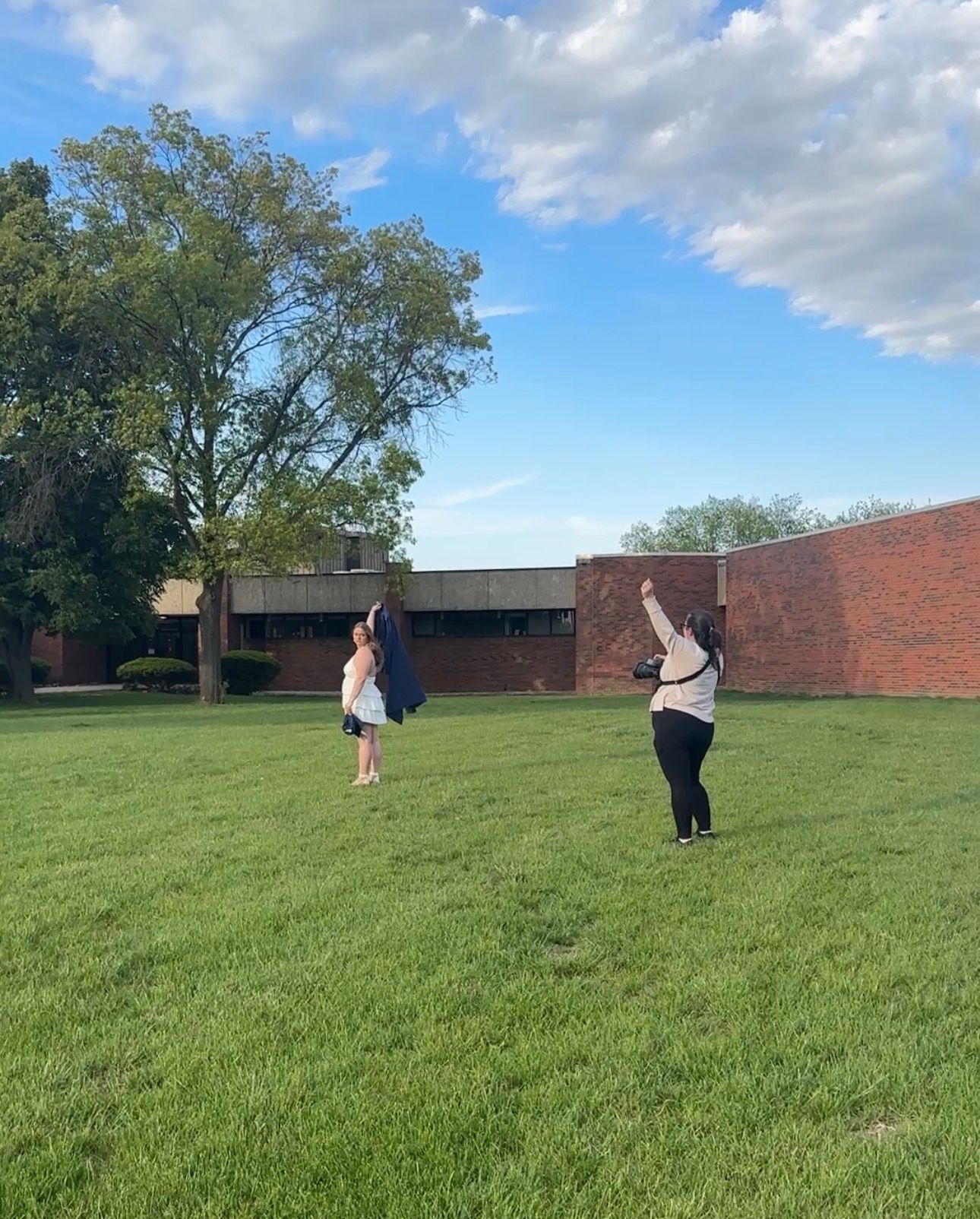 Two people playing outside on a green lawn near a brick school building under a partly cloudy blue sky.