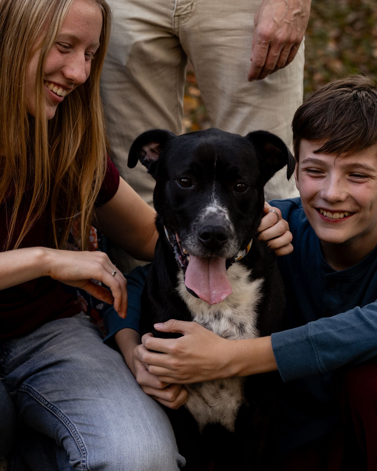 Two smiling children sit close together outdoors hugging a happy black and white dog with its pink tongue out while an adult stands behind.