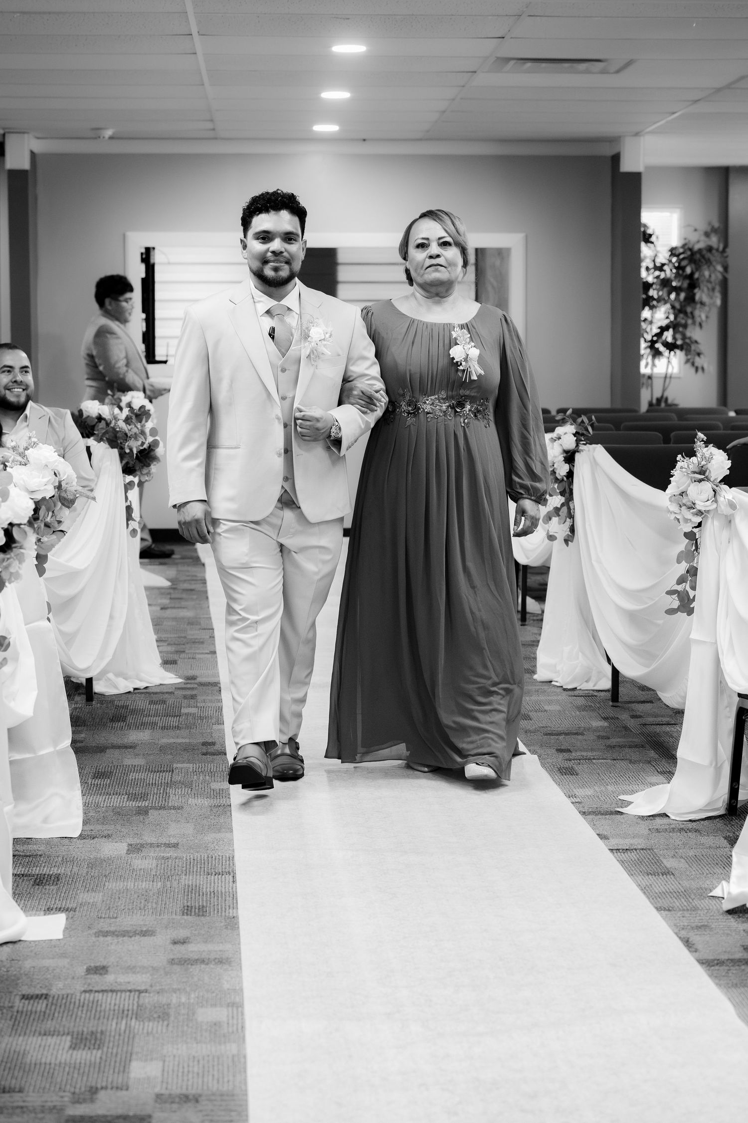 A groom in a light suit escorts a woman in a long dark dress down a wedding aisle lined with floral pew decorations.