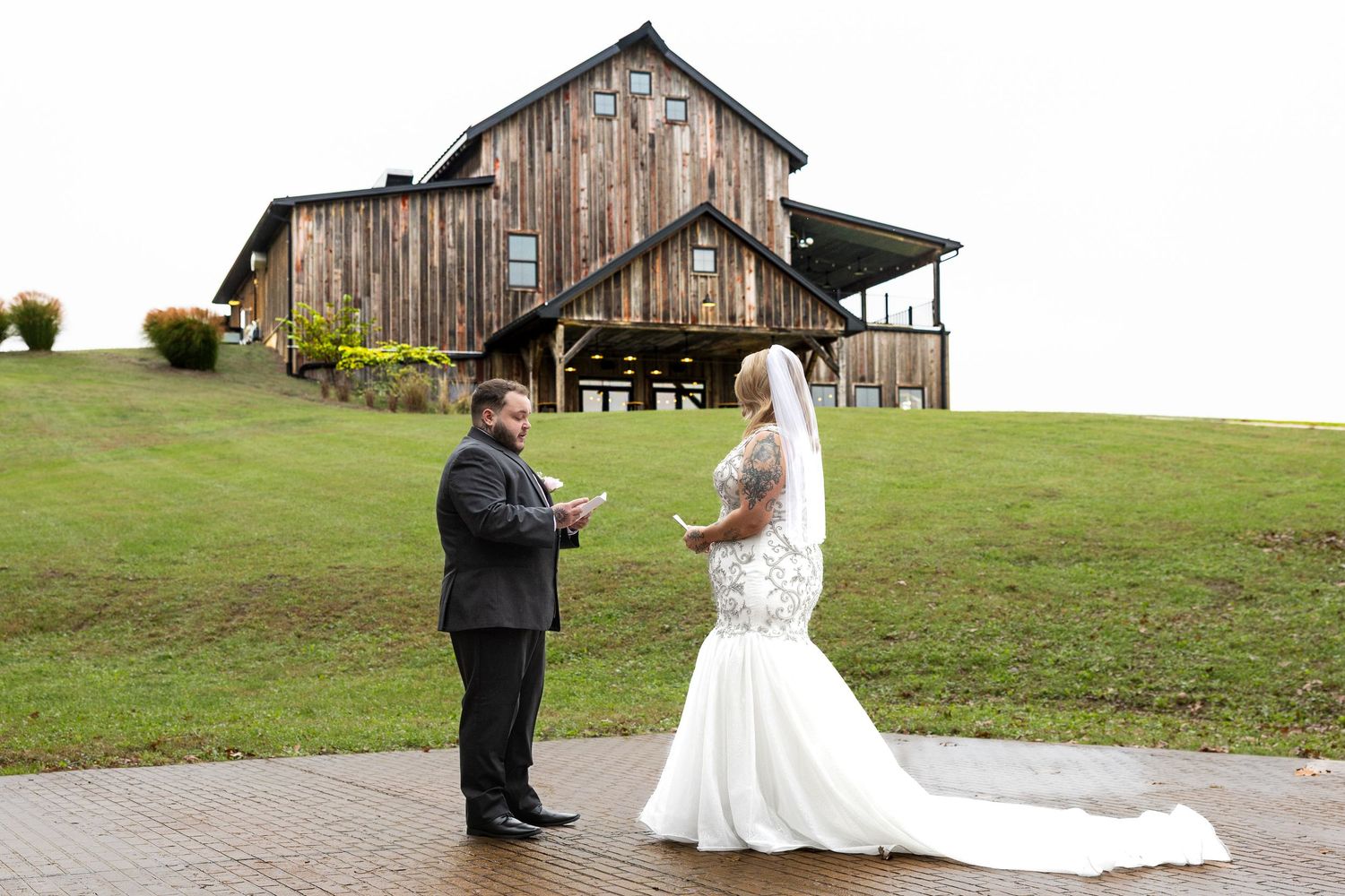 Bride in a lace wedding gown and groom in a dark suit read vows outdoors in front of a rustic wooden barn venue on a green hillside.