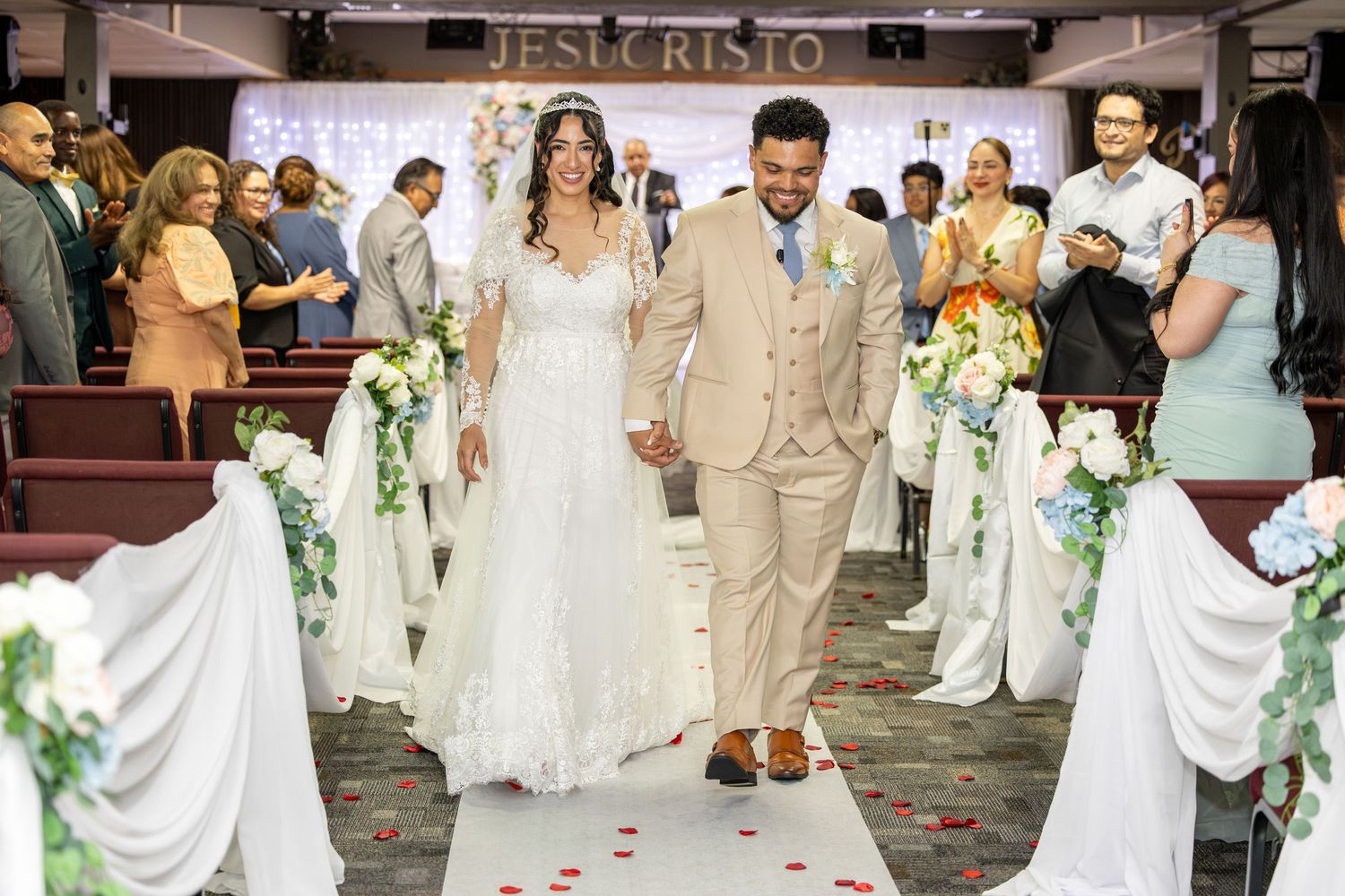 Bride in a white gown and groom in a tan suit walk hand in hand down a petal-strewn aisle lined with white flowers as wedding guests watch.