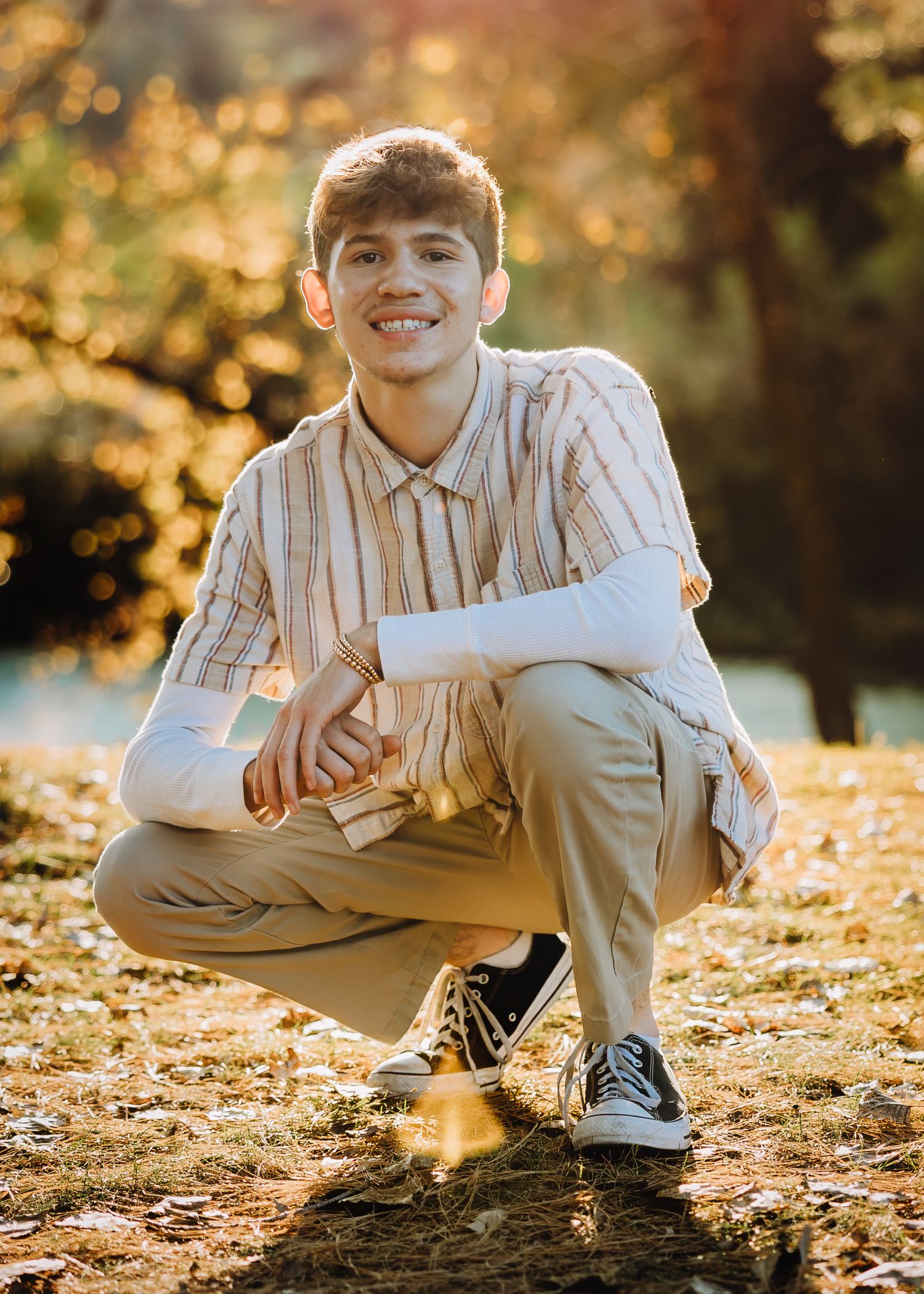Smiling teenage boy crouching outdoors in autumn park, wearing striped shirt and khaki pants with Converse sneakers.
