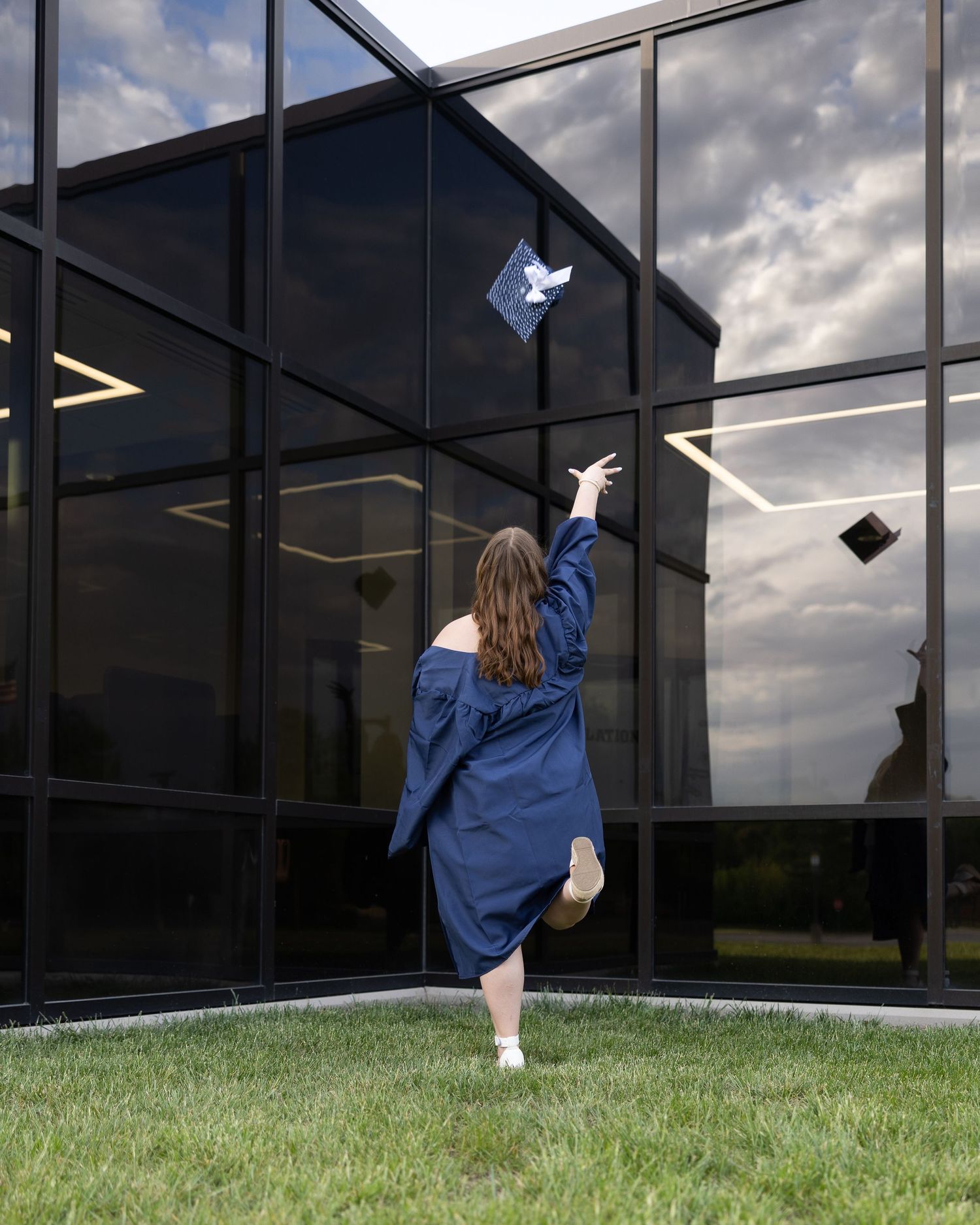 Graduate in navy gown tossing cap in air outside modern glass building on green lawn.