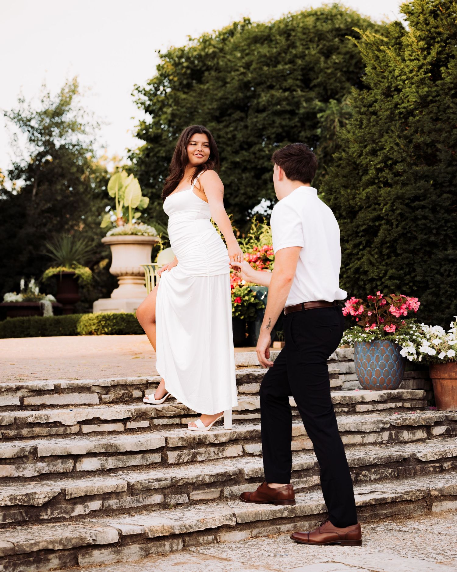 Couple in white outfits on stone steps in a garden, woman smiling down at man holding flowers.