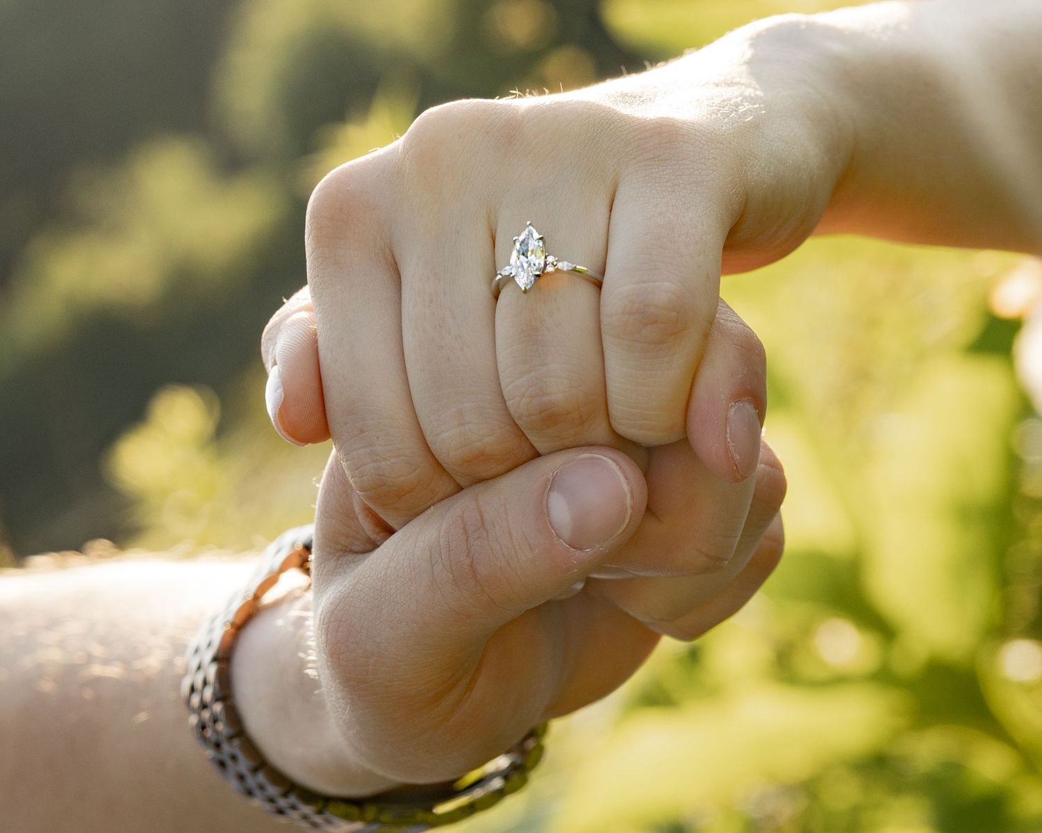 Close-up of two hands holding, woman wearing a marquise diamond solitaire engagement ring, green nature background.