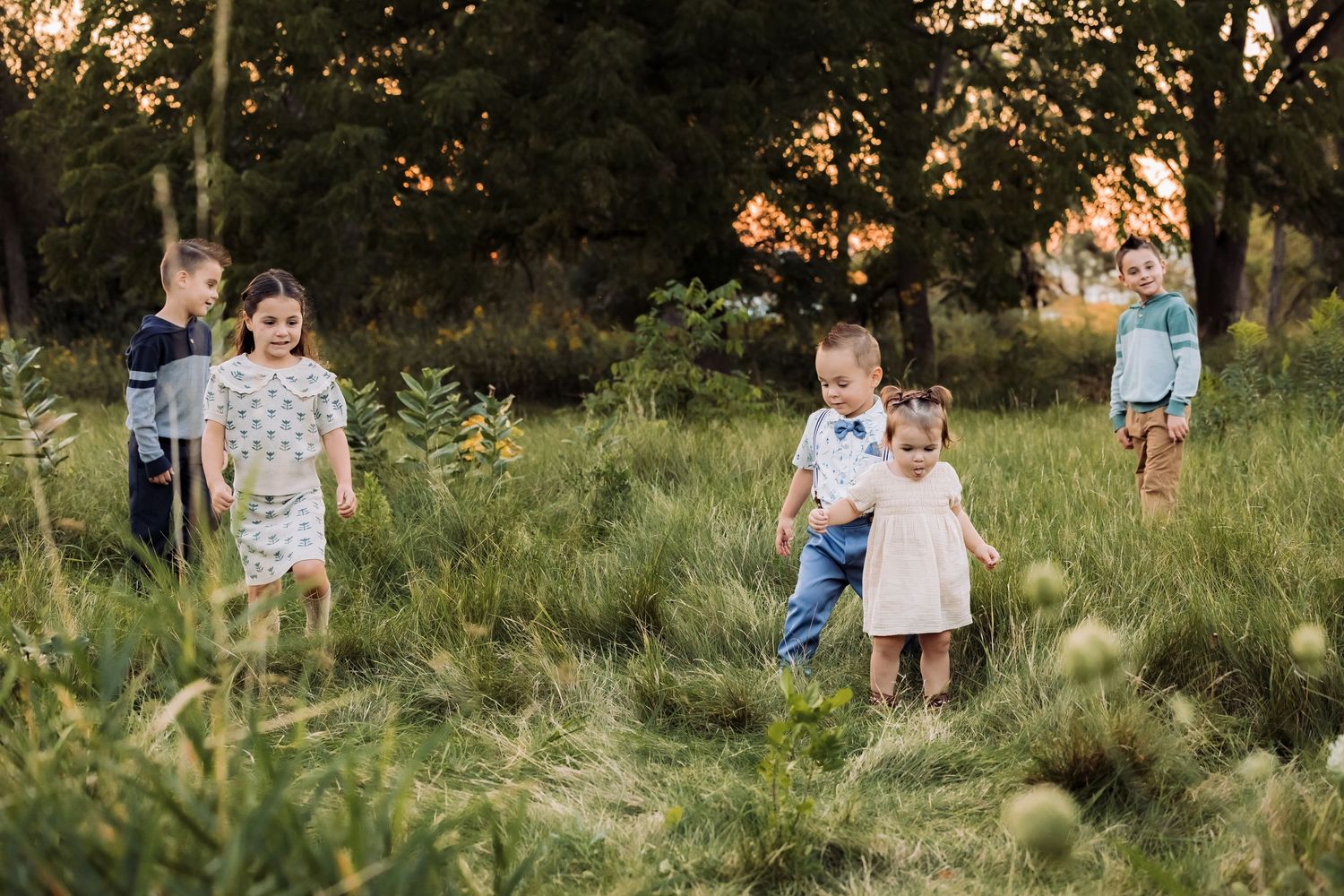 Five young children playing in a lush green meadow at golden hour sunset with trees in background.