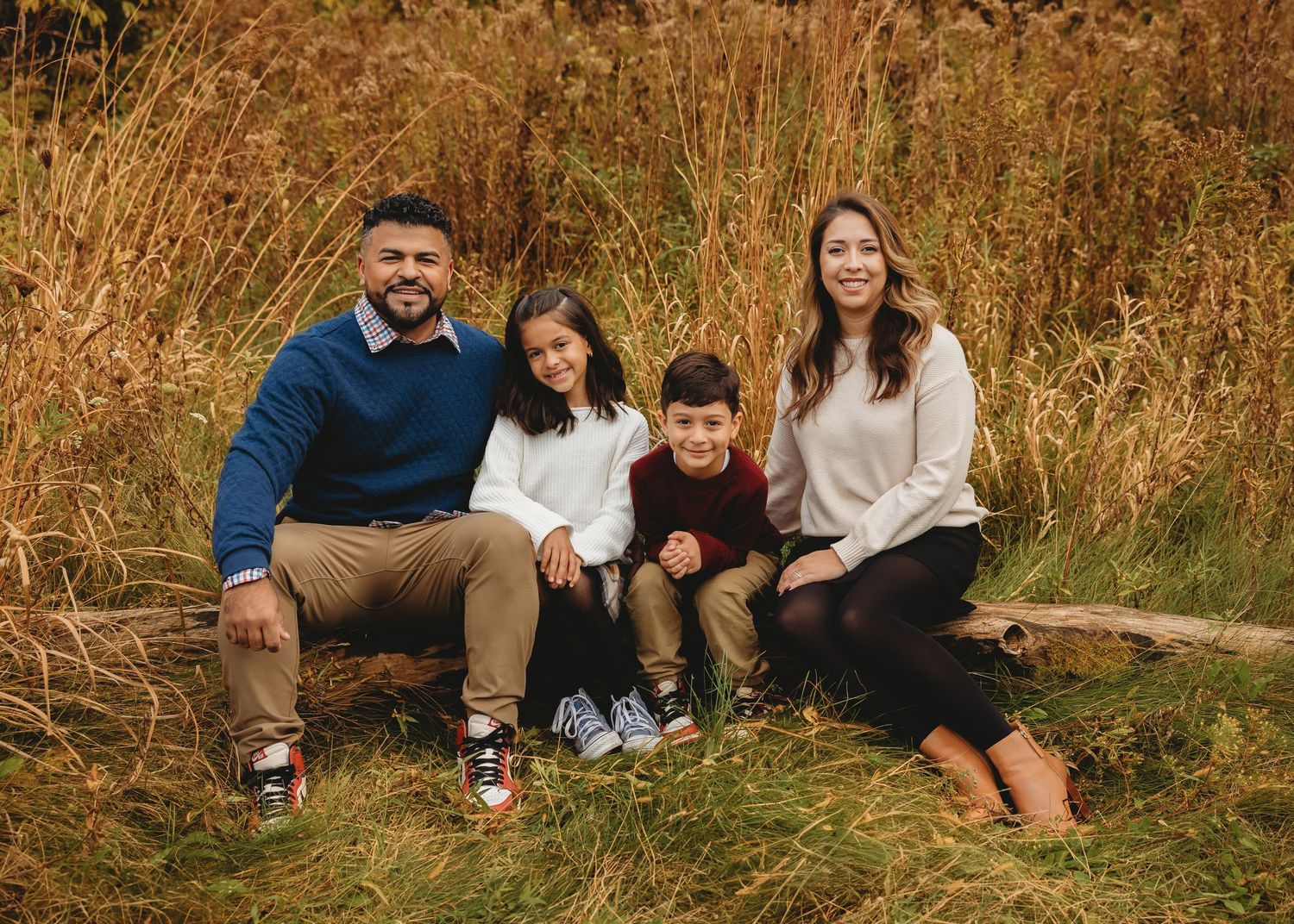 Happy family of four sitting together in golden autumn grass field, smiling at the camera.