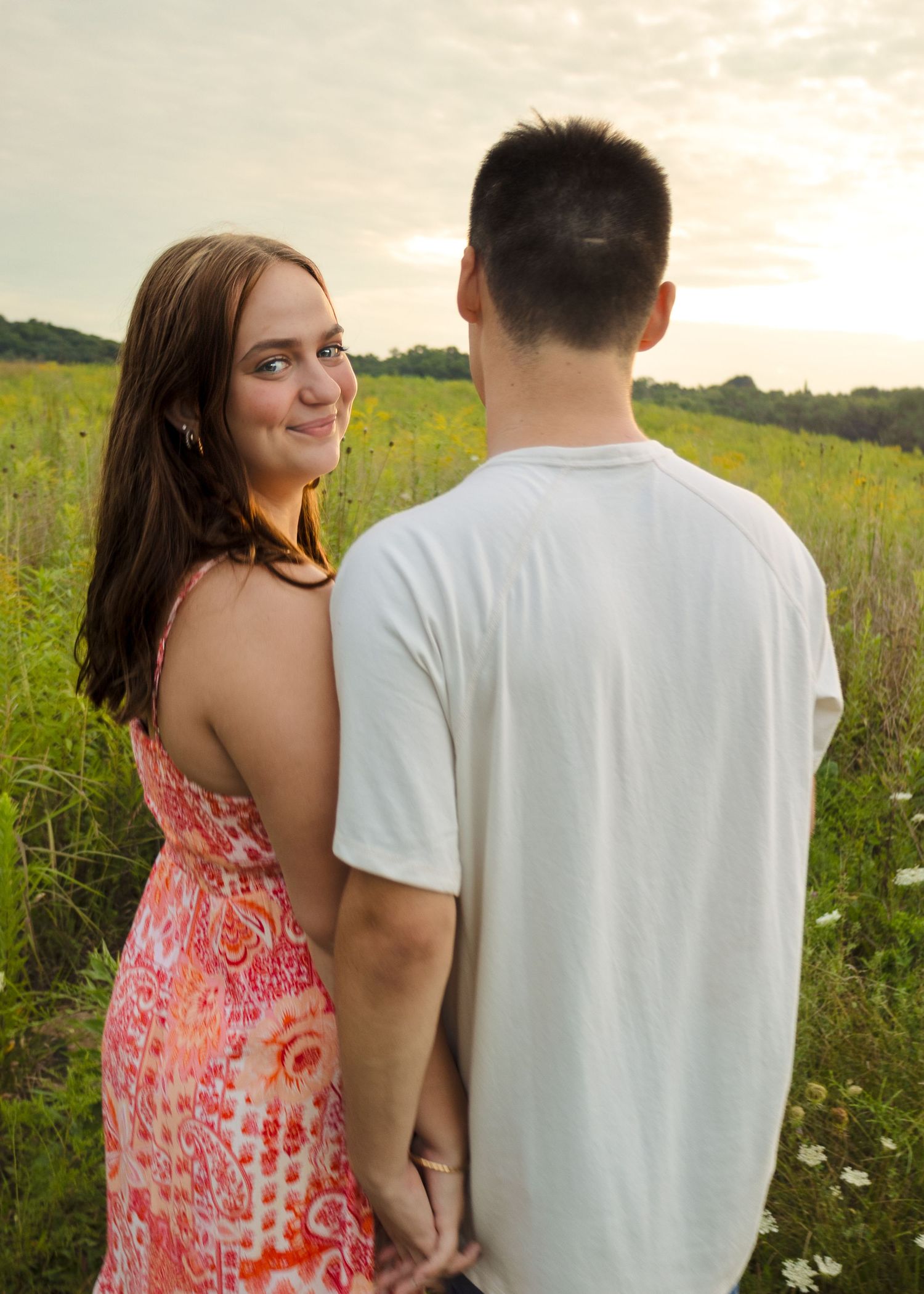 Young woman in pink floral dress smiling over shoulder while holding hands with man in white tee in a field.