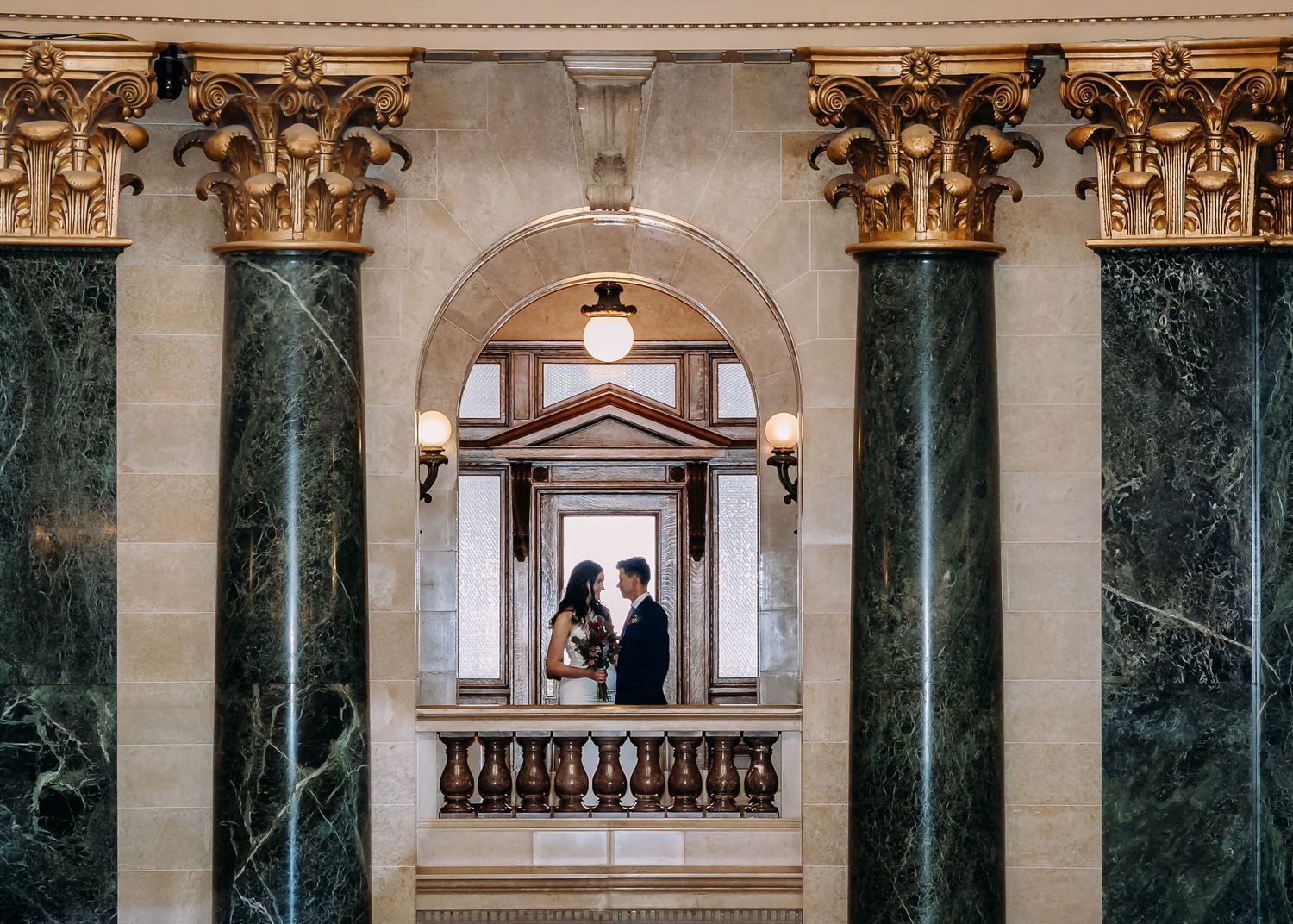 Wedding couple embracing between towering green marble columns under a decorative stone arch.