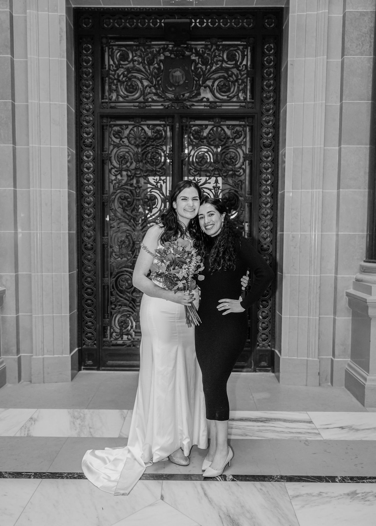 Black and white photo of bride in fitted gown embracing guest before ornate carved wooden doors.