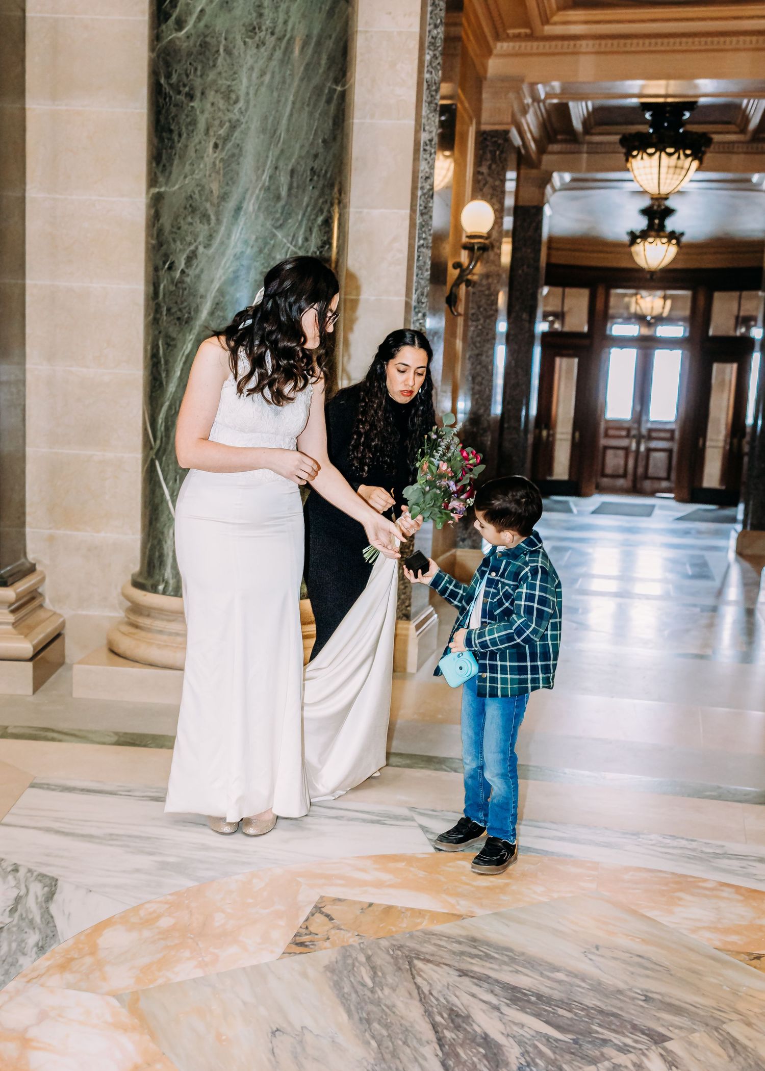 Bride in white dress and female guest accepting a flower bouquet from a young boy in a marble hallway.