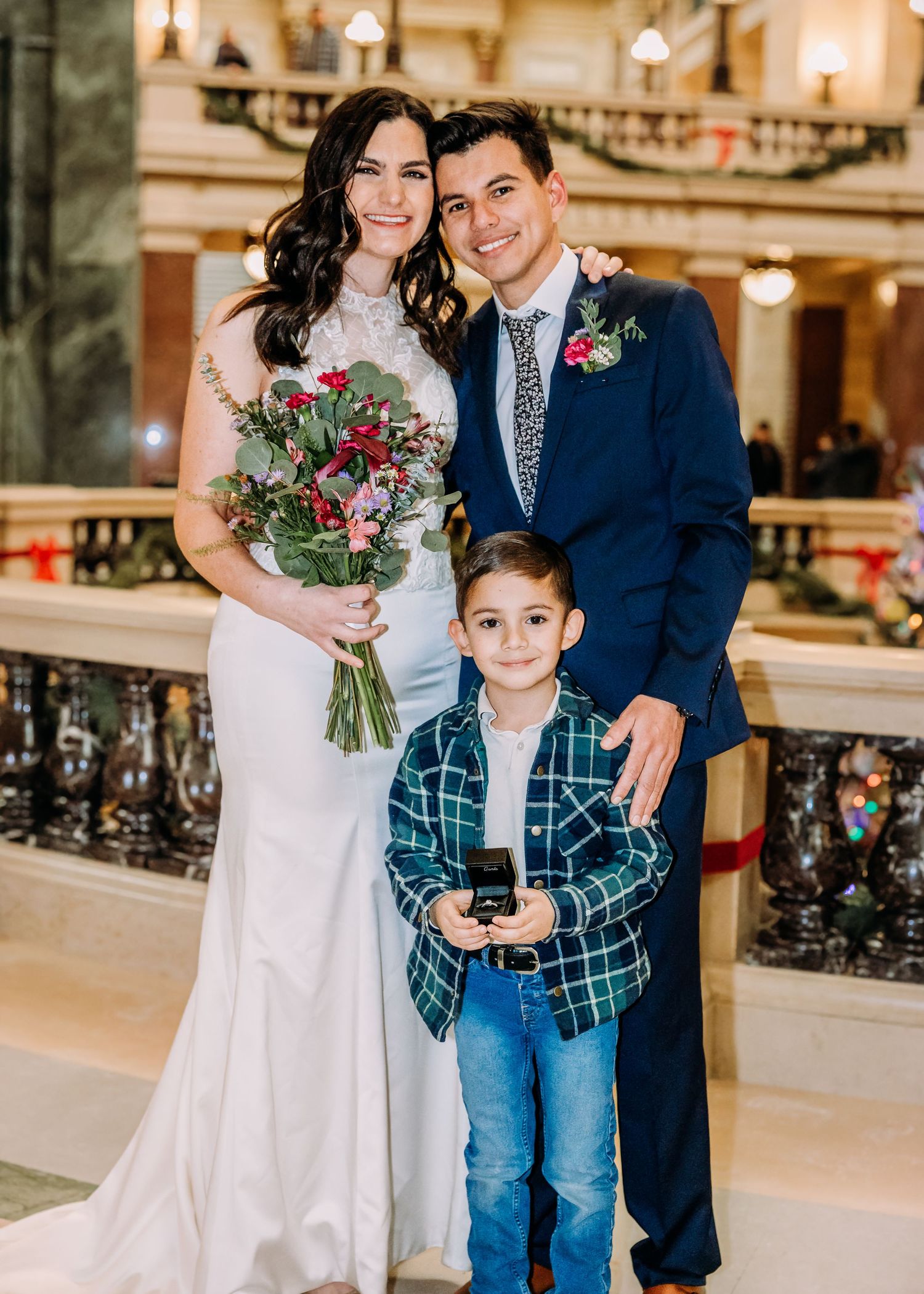 Happy wedding couple in navy suit and white gown poses with young boy in plaid at ceremony.