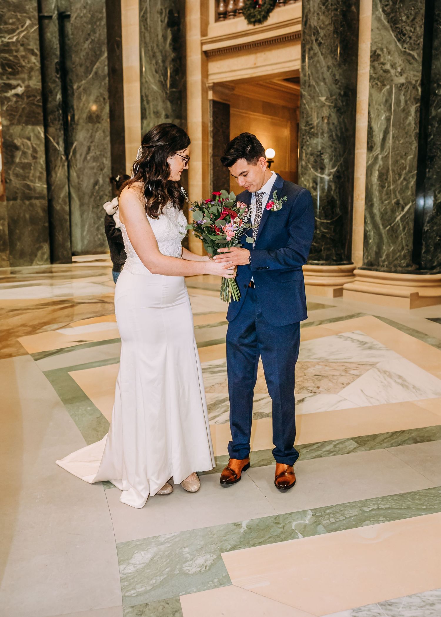 Bride in white gown and groom in blue suit sharing a bouquet in a grand marble wedding venue.
