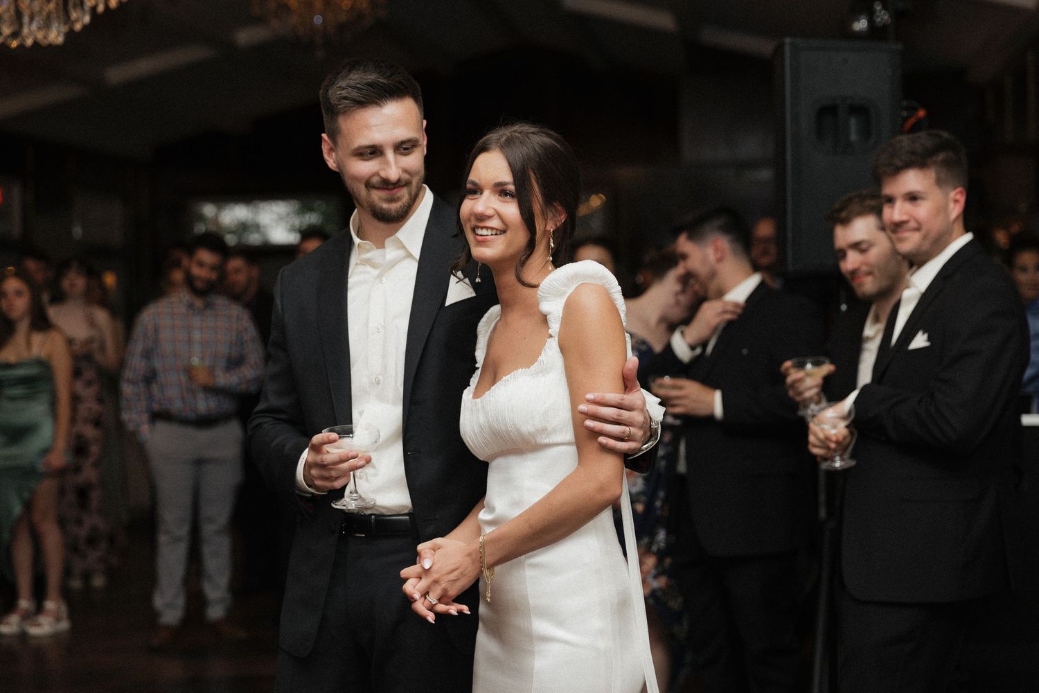 A couple celebrates during their wedding reception as guests gather in the background at a romantically lit venue.