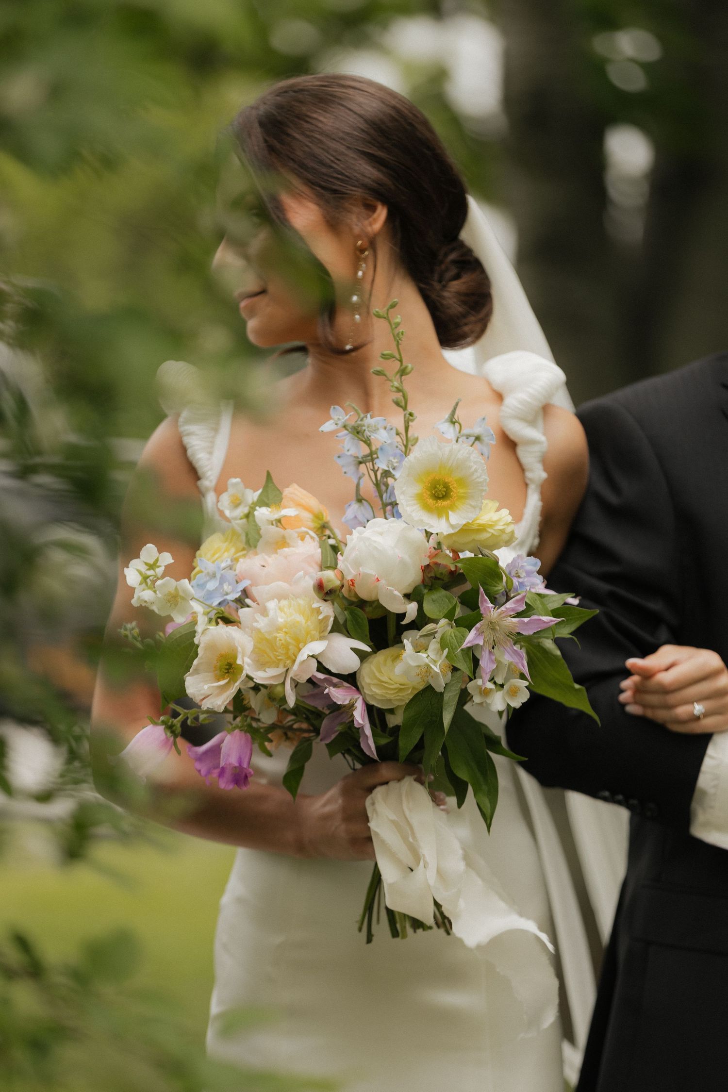A stunning bridal bouquet featuring yellow, white and purple flowers is held against an elegant white wedding dress.
