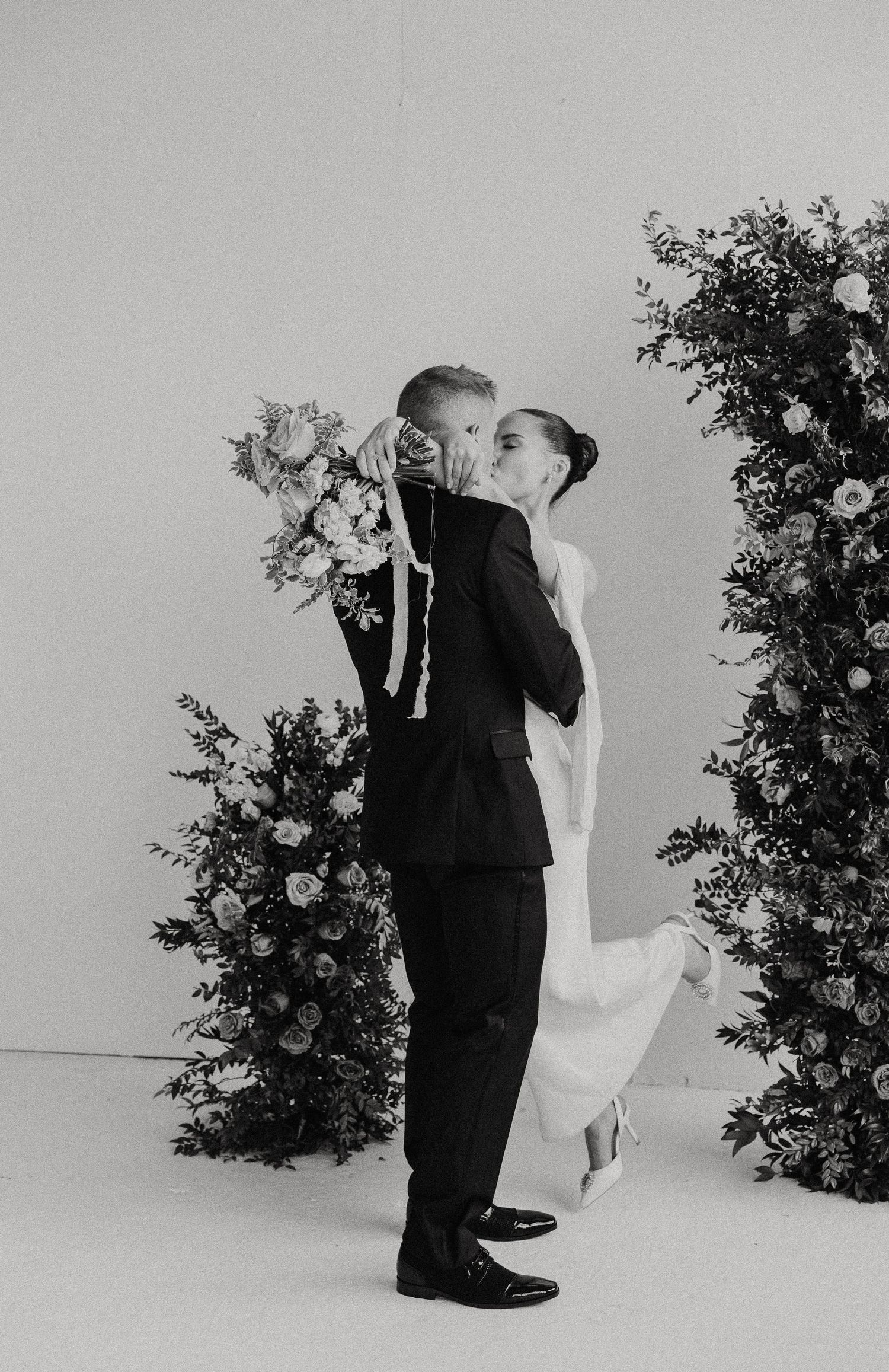 A black and white photo of a couple embracing among floral arrangements.