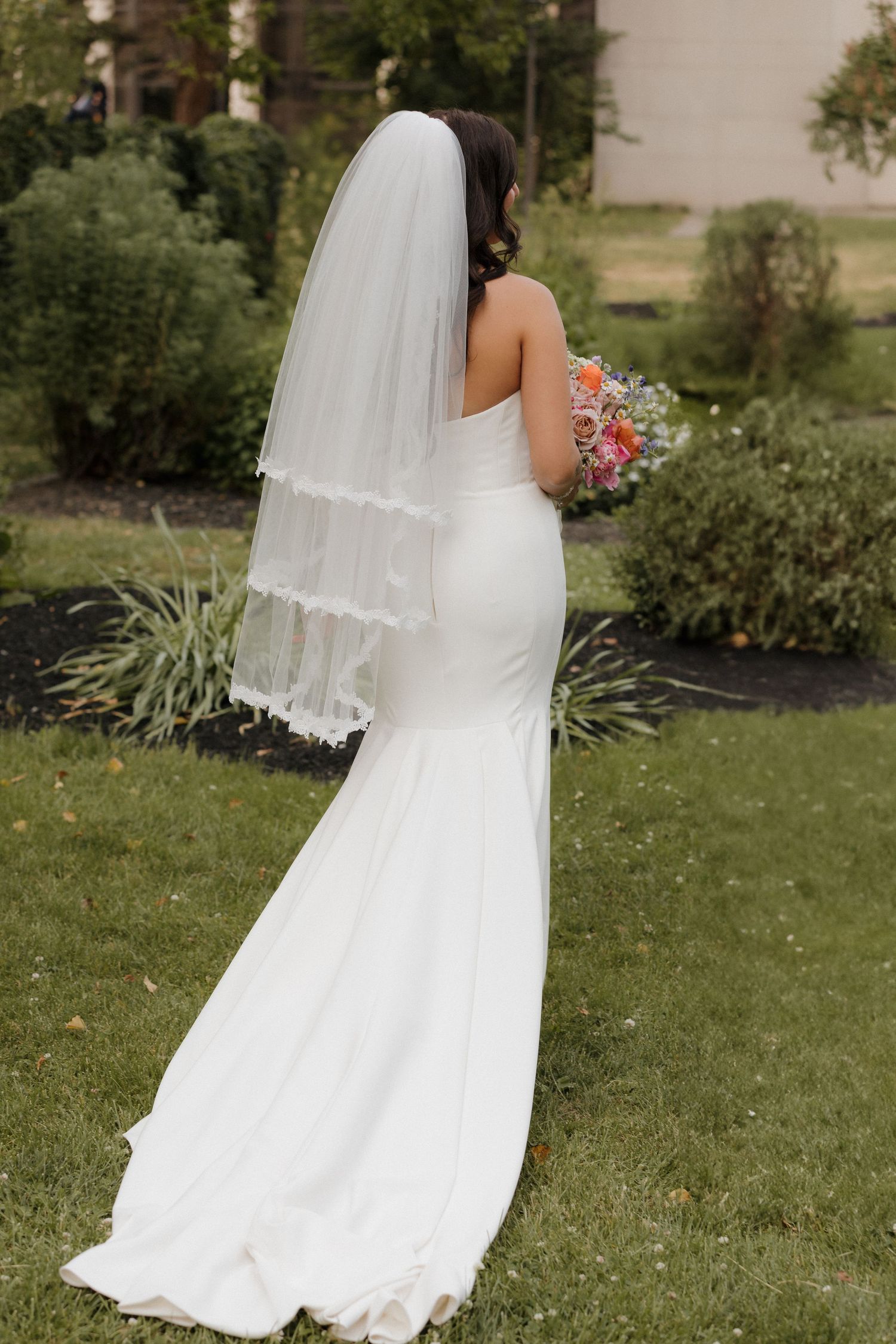 A bride in a fitted white mermaid wedding gown and veil holds a coral bouquet while standing on grass.