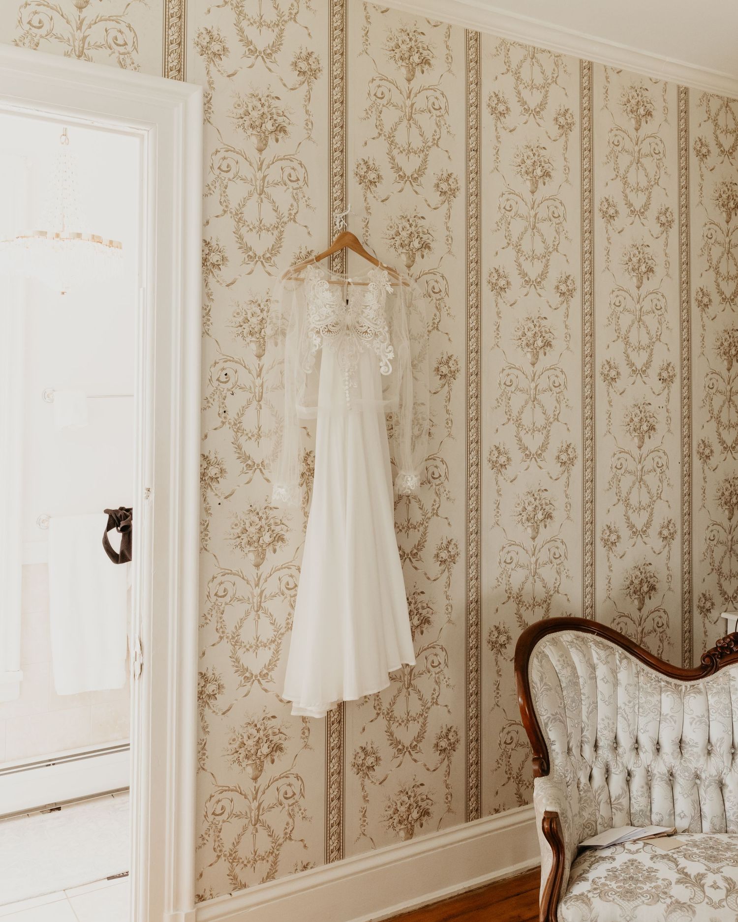 A vintage wedding dress hangs on a wooden hanger against vintage floral wallpaper next to an antique chair. maison albion wedding venue.