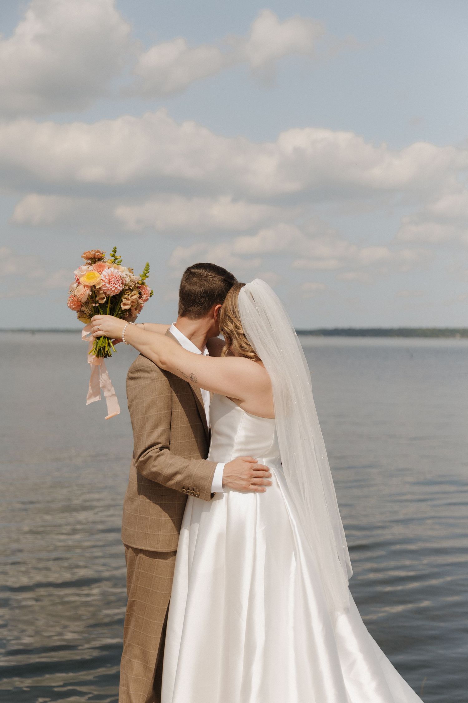 A couple embraces by a lake with the bride holding a colorful bouquet against a cloudy sky.