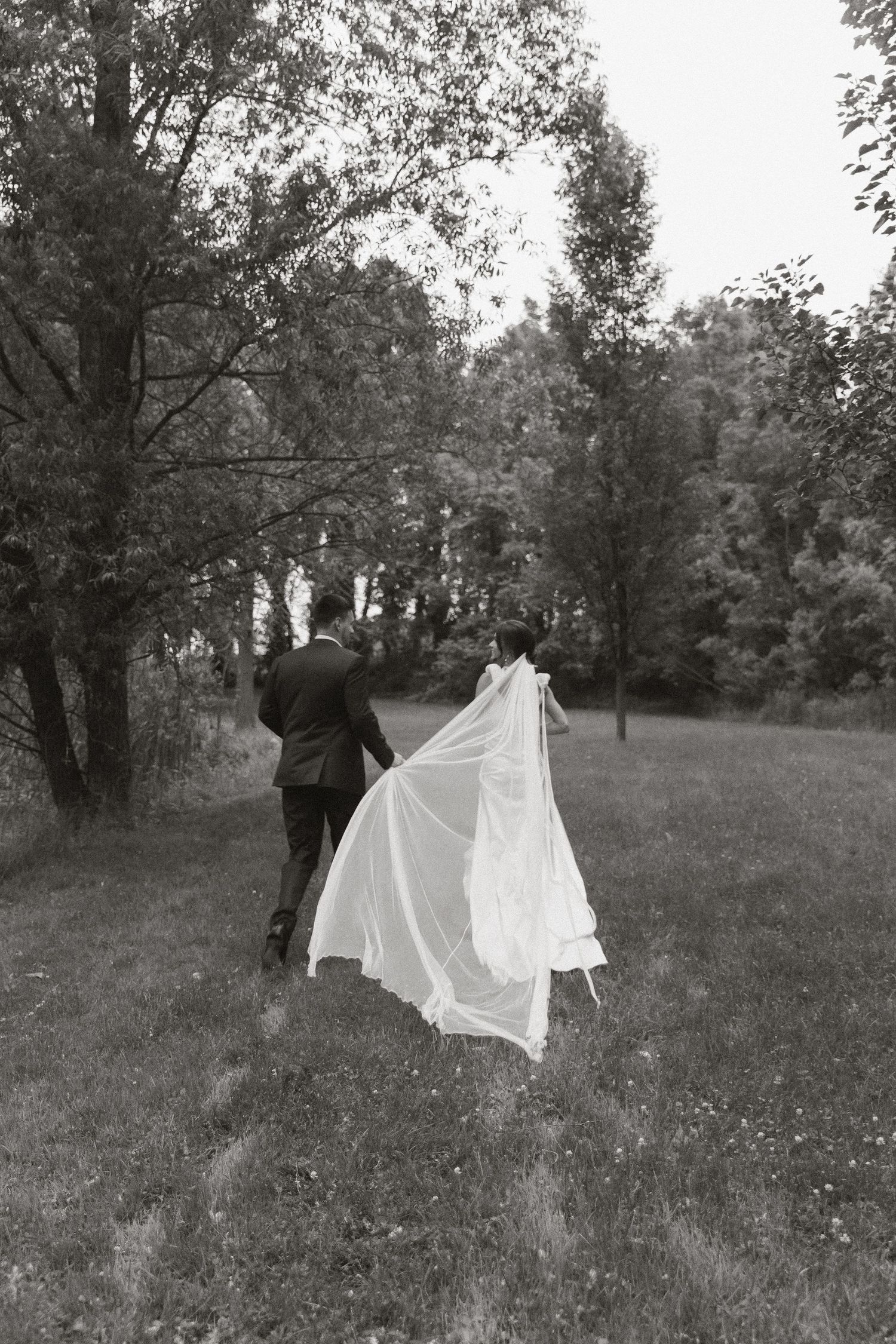 Black and white image of wedding couple walking through a meadow with flowing dress.