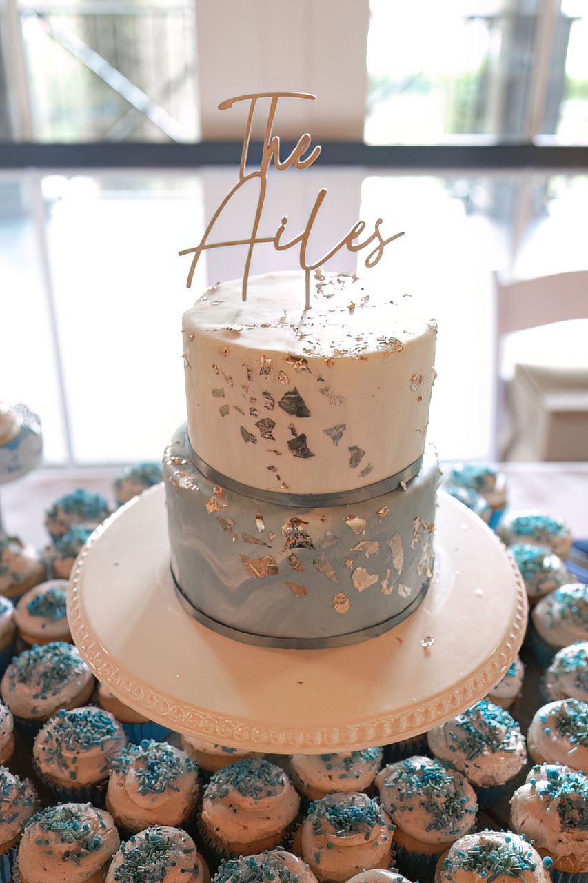 Wedding cake with silver accents and blue cupcakes arranged on white stand.
