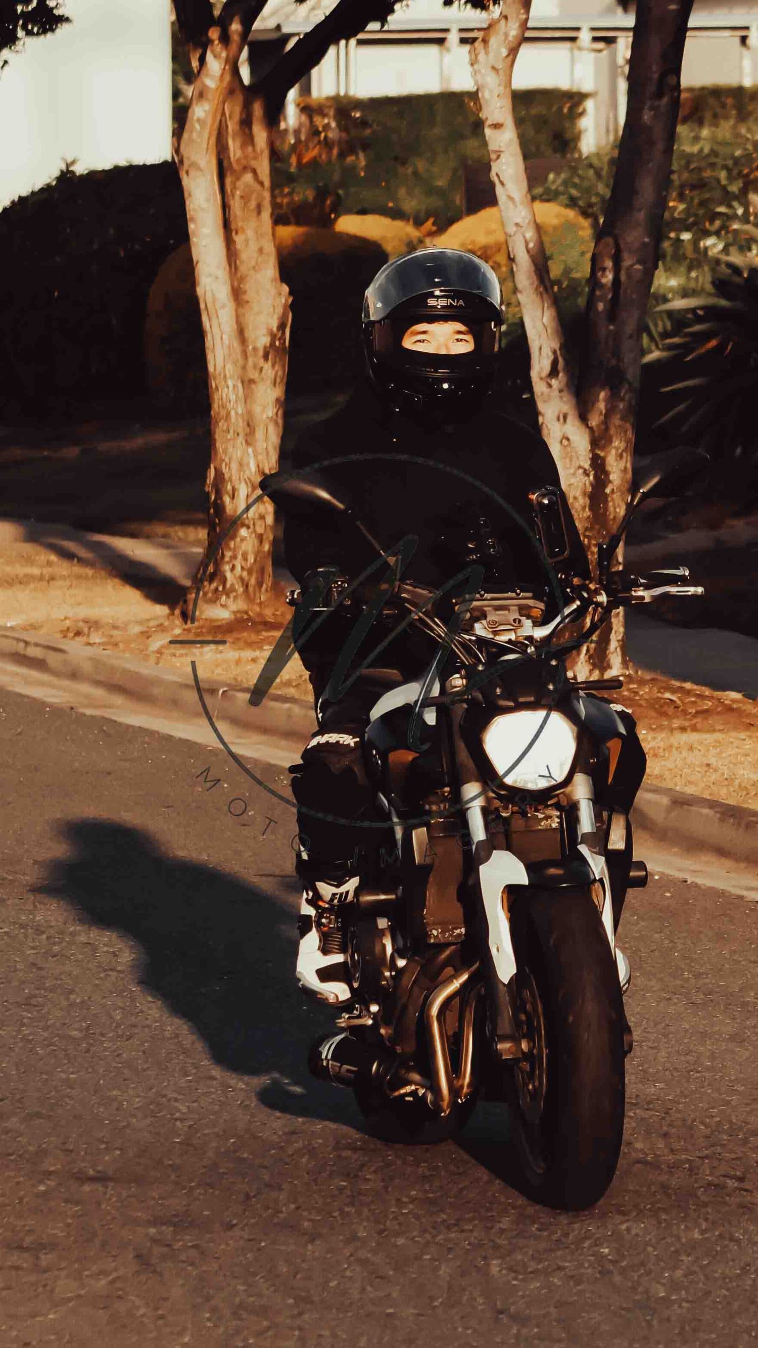 A motorcyclist in black gear and helmet rides a motorcycle on a street lined with trees.