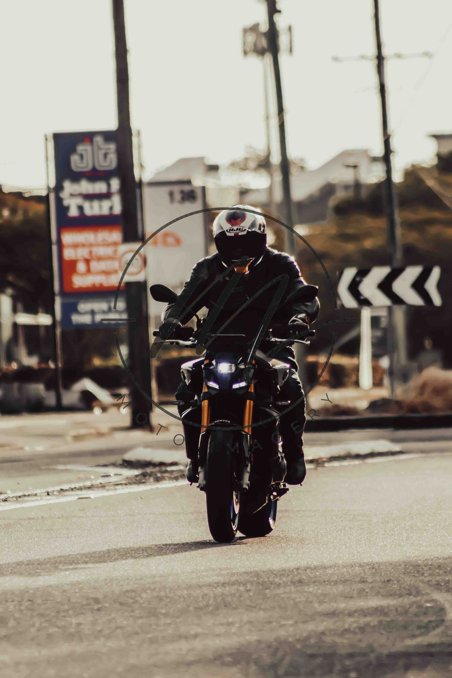 Motorcyclist in black leather jacket and helmet riding a naked motorcycle on a road near a John T Turf wholesale store.