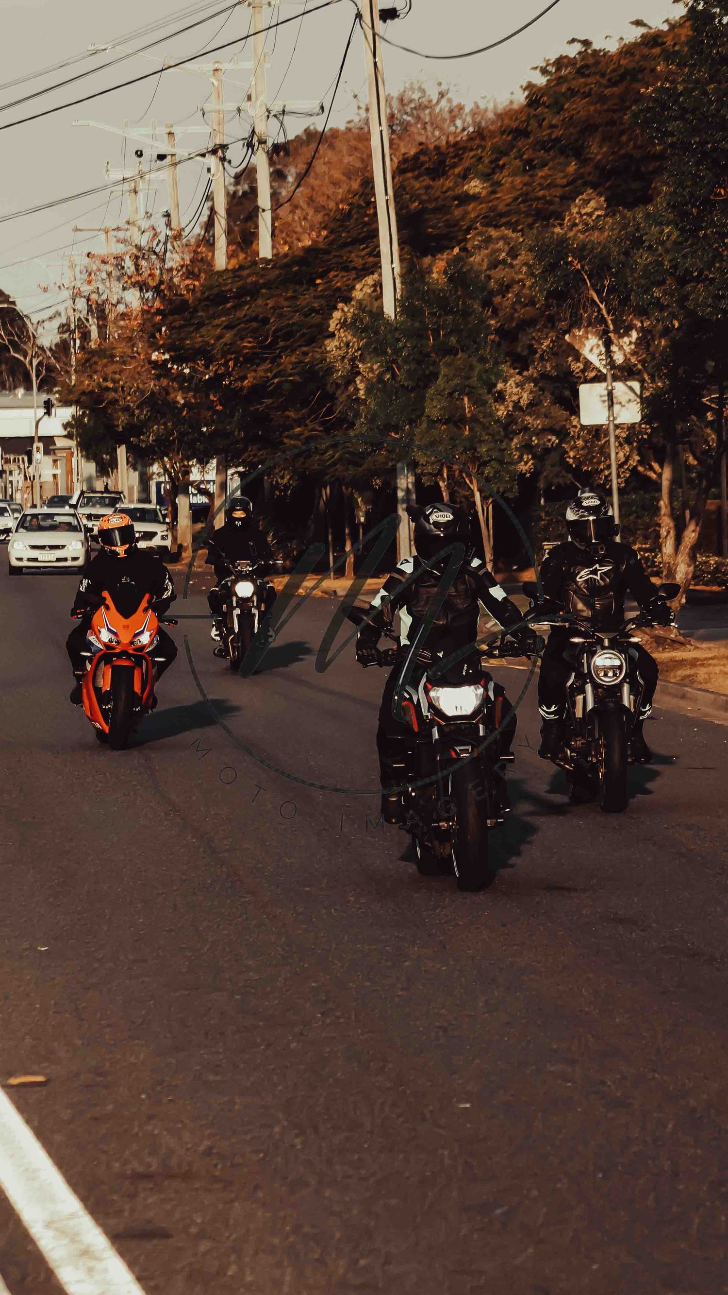 Group of motorcyclists riding in formation on a tree-lined urban street, featuring a bright orange sport bike in front.