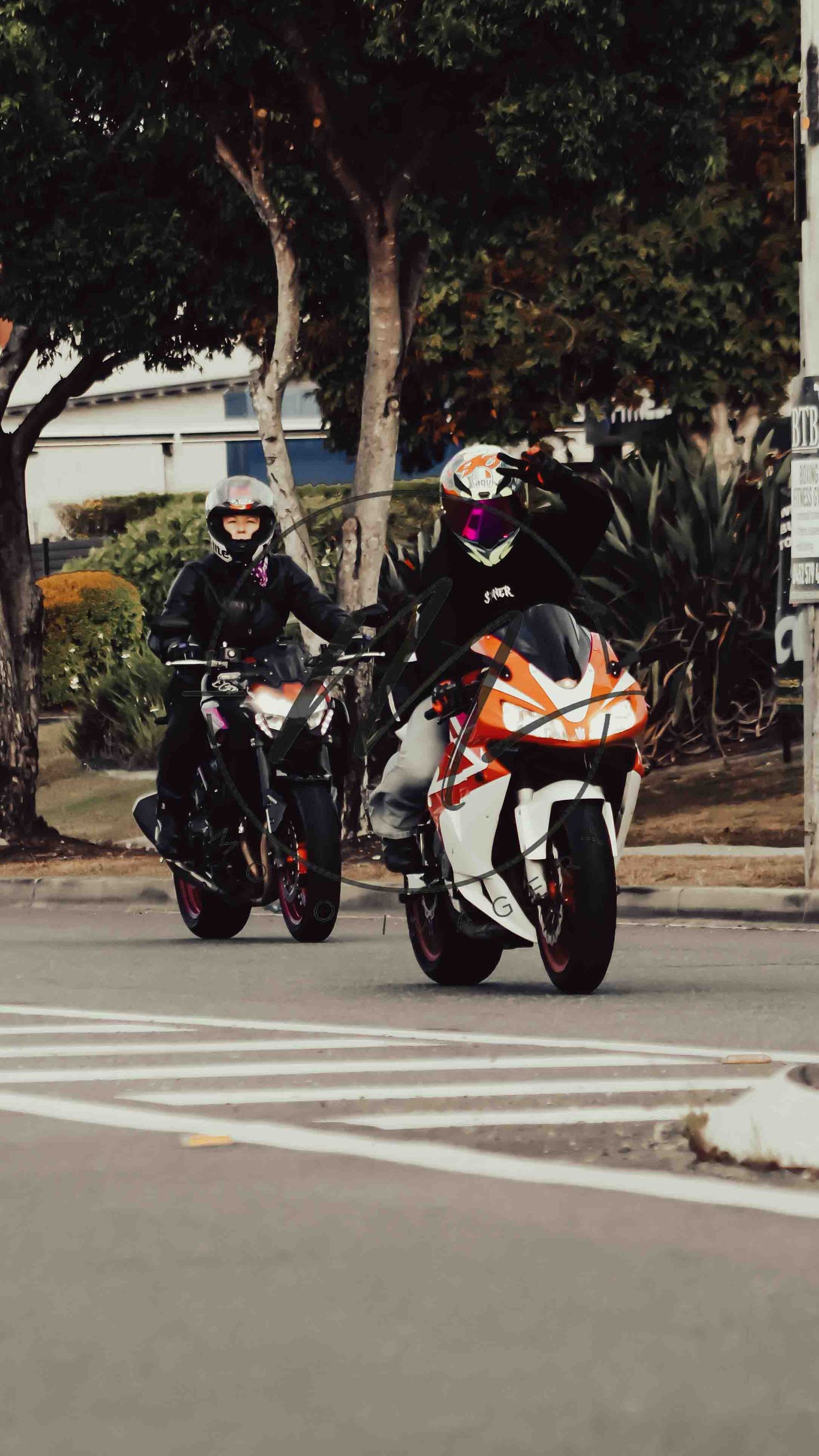 Two motorcyclists in helmets riding KTM bikes down a suburban street lined with trees.
