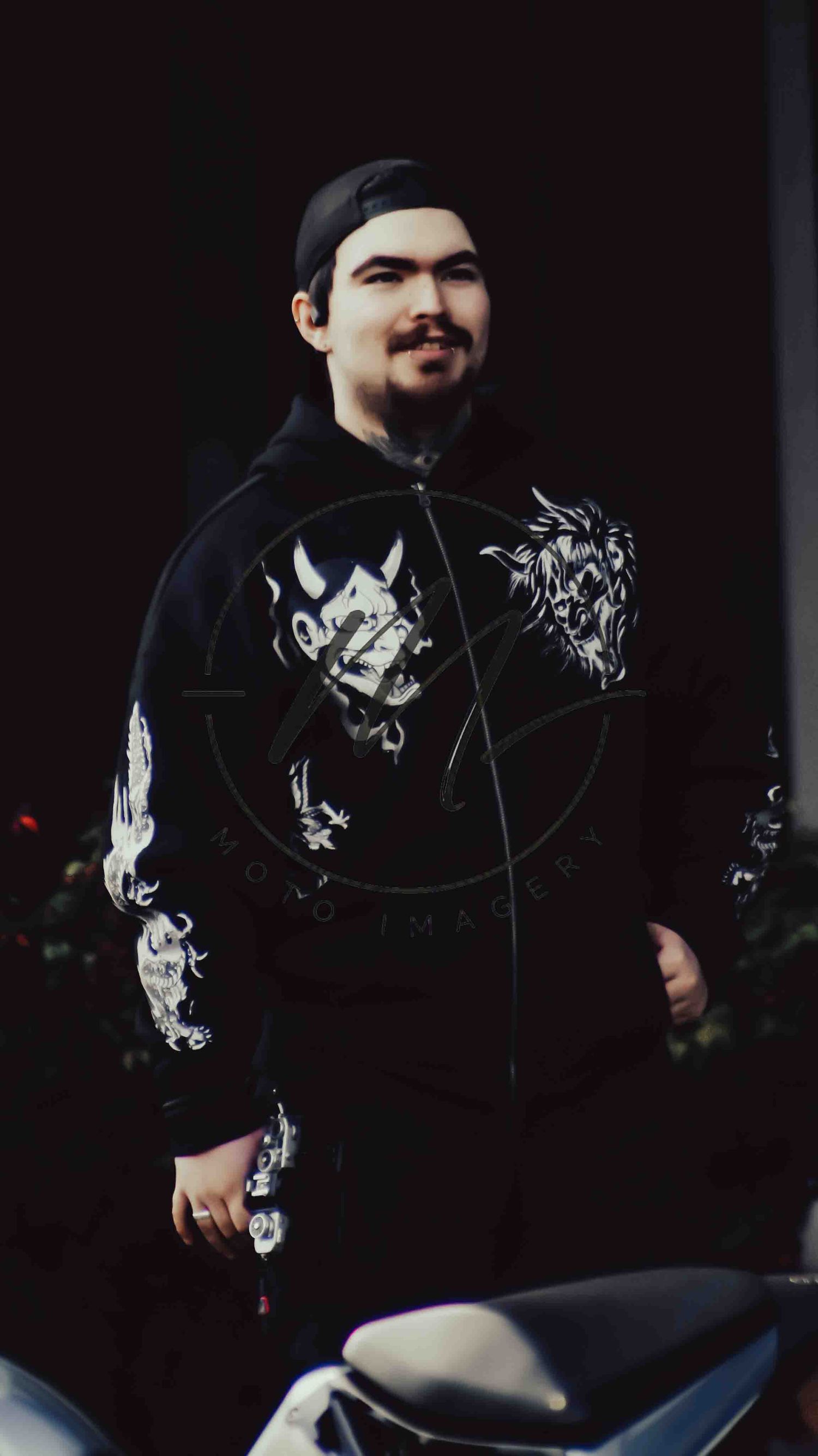 Young man smiling in black dragon-print jacket and cap, posed confidently in a dark atmospheric setting.