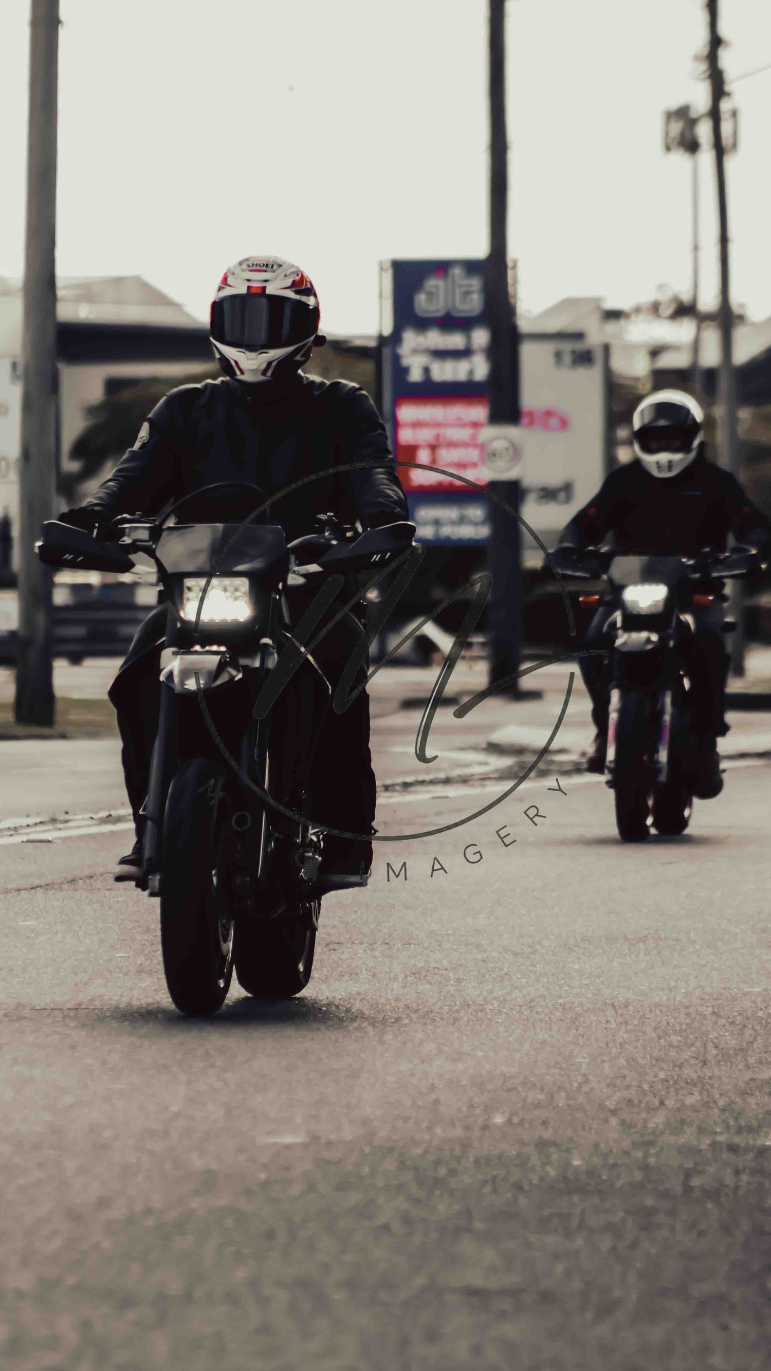 Two motorcyclists in black gear and helmets riding on a city street, captured in a moody, dark-toned photograph.