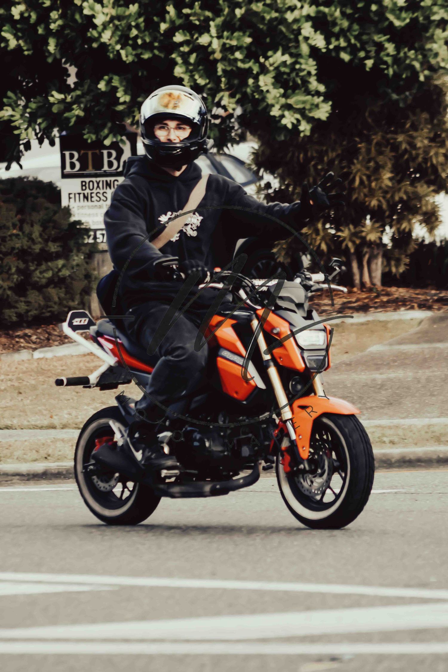 Rider on orange Honda Grom mini motorcycle wearing black hoodie and white helmet on city street.