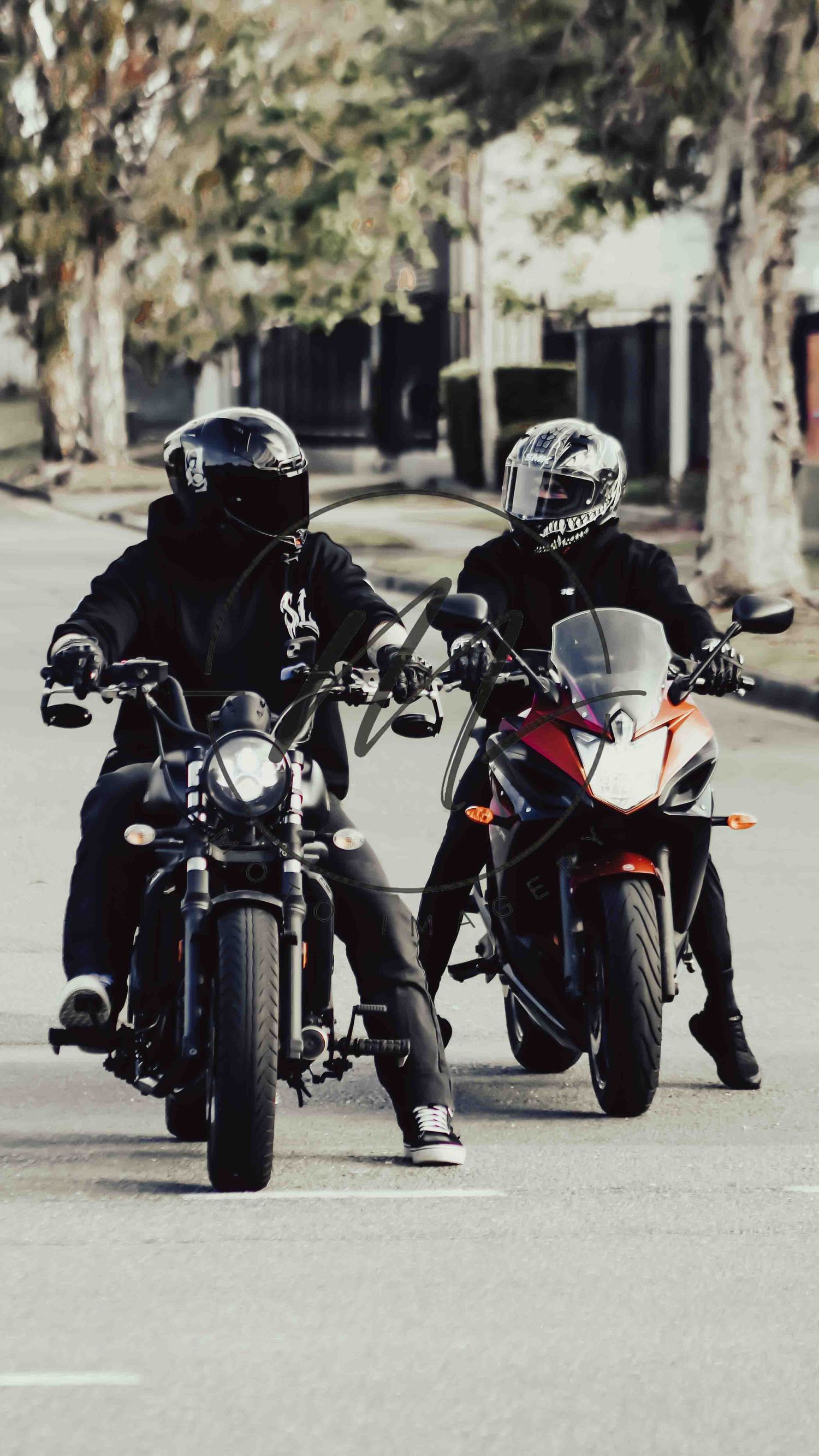Two motorcyclists in black jackets and helmets riding sport bikes side by side on a tree-lined street.