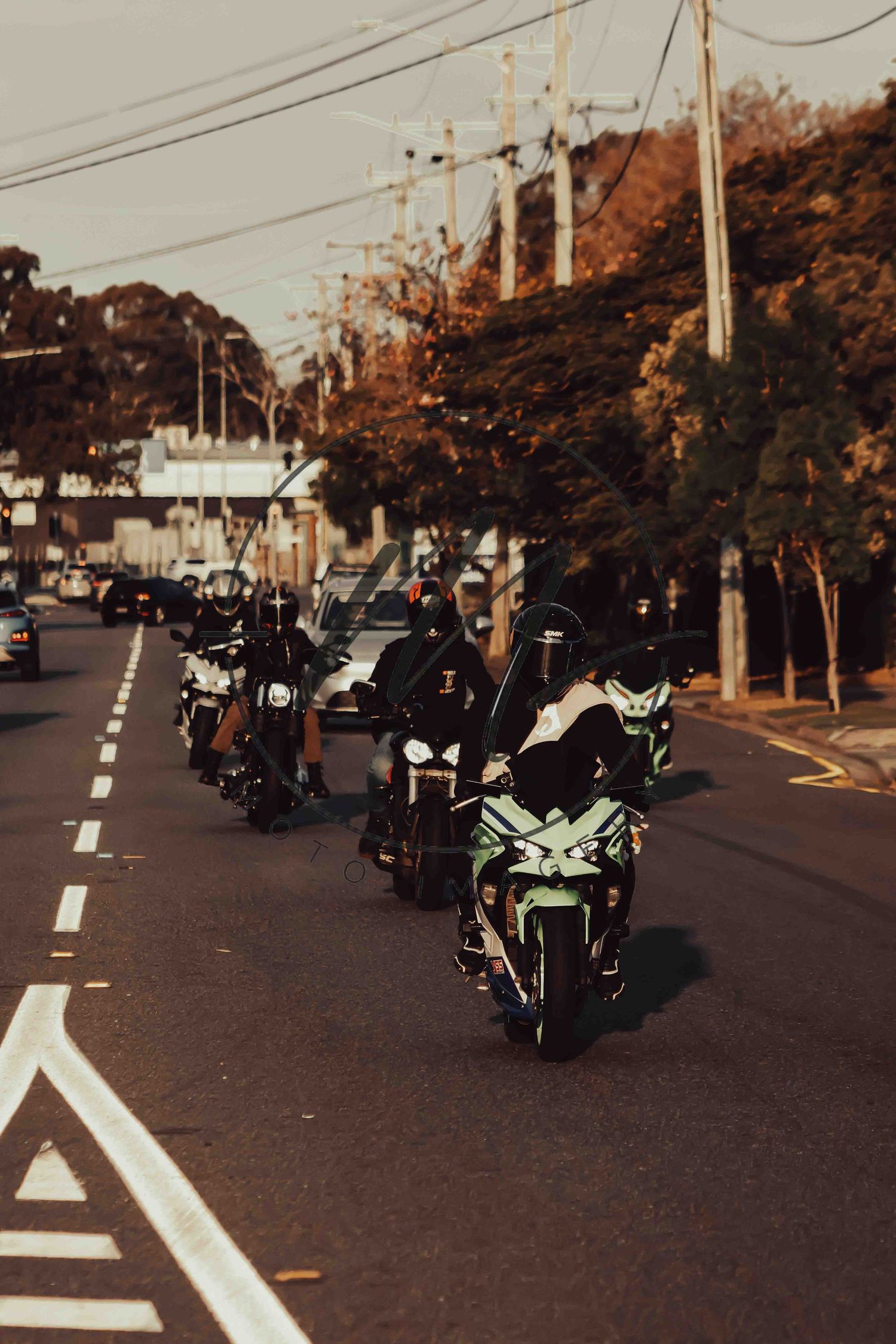 A group of motorcyclists riding in formation down a urban street lined with trees and power lines.