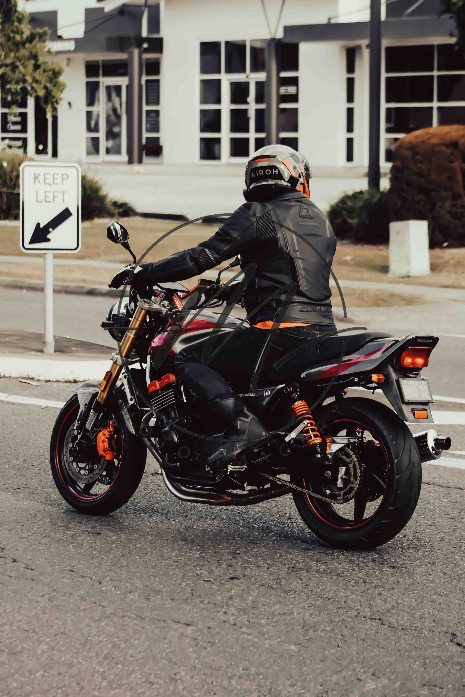 Motorcyclist in black leather jacket and helmet riding a sport motorcycle past a Keep Left sign on a city street.