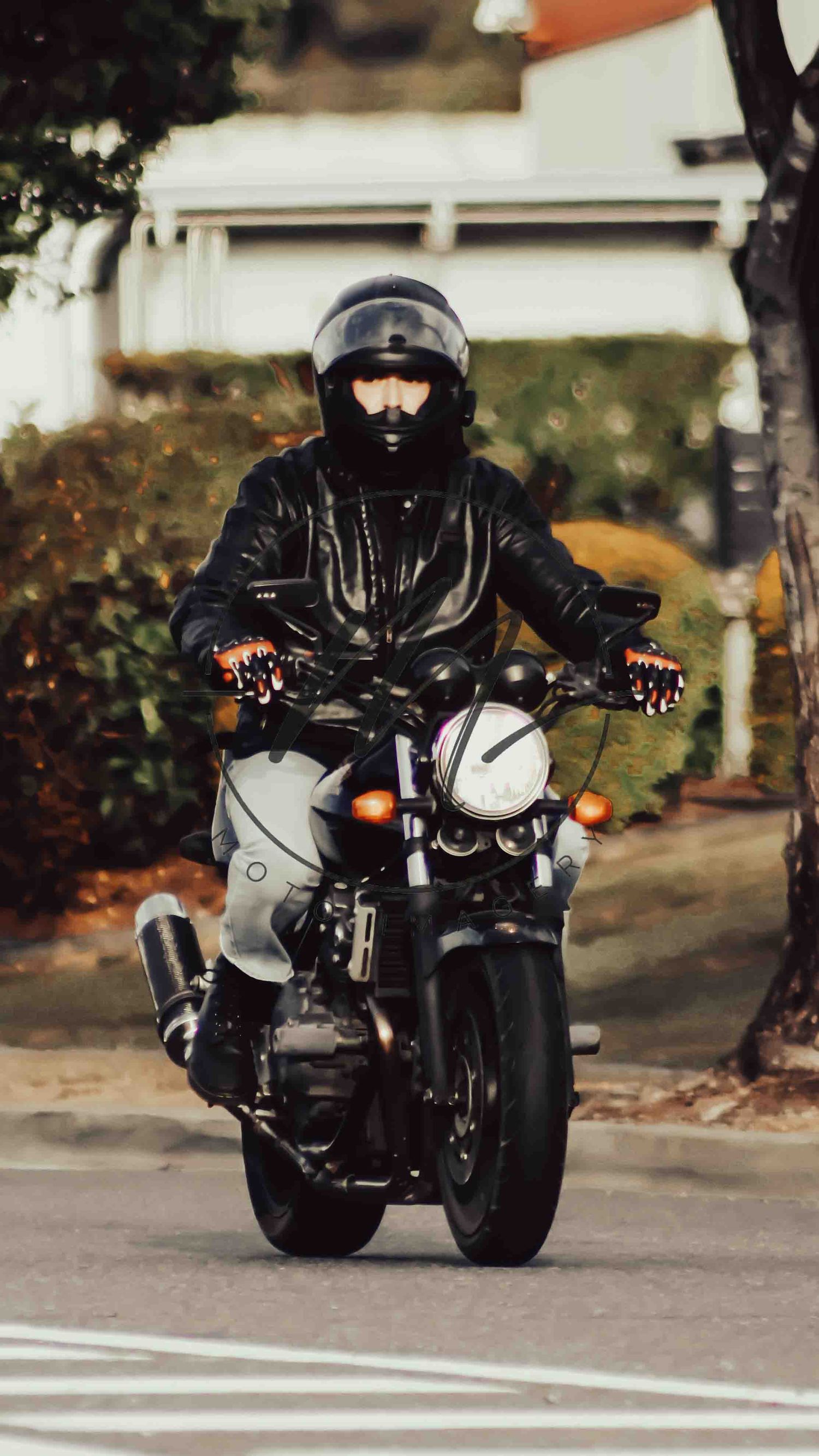 Motorcyclist in black leather jacket and helmet riding a classic motorcycle on a suburban street.