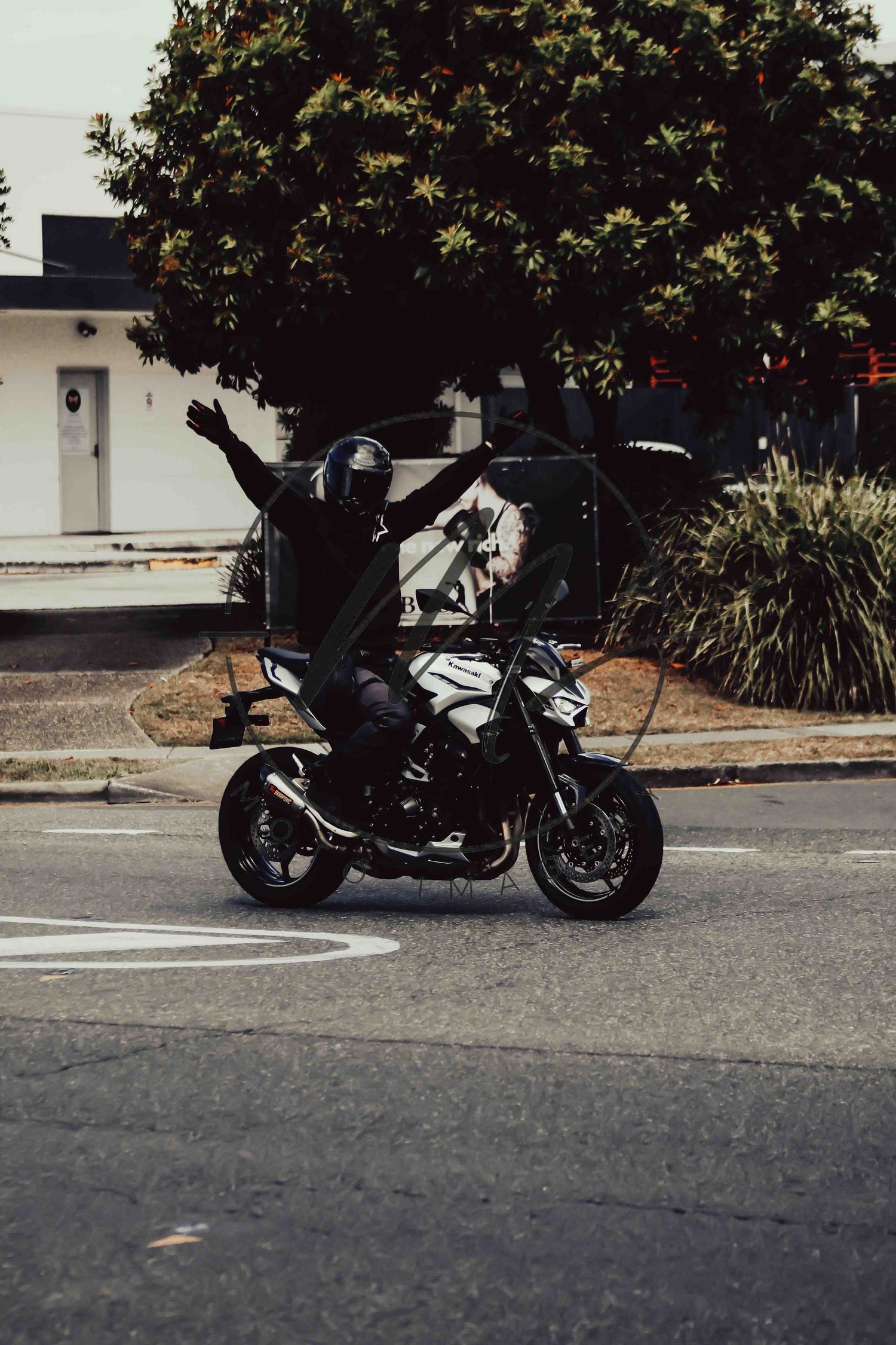 Motorcyclist with arms raised riding a black sport bike on a street corner with trees and buildings in background.