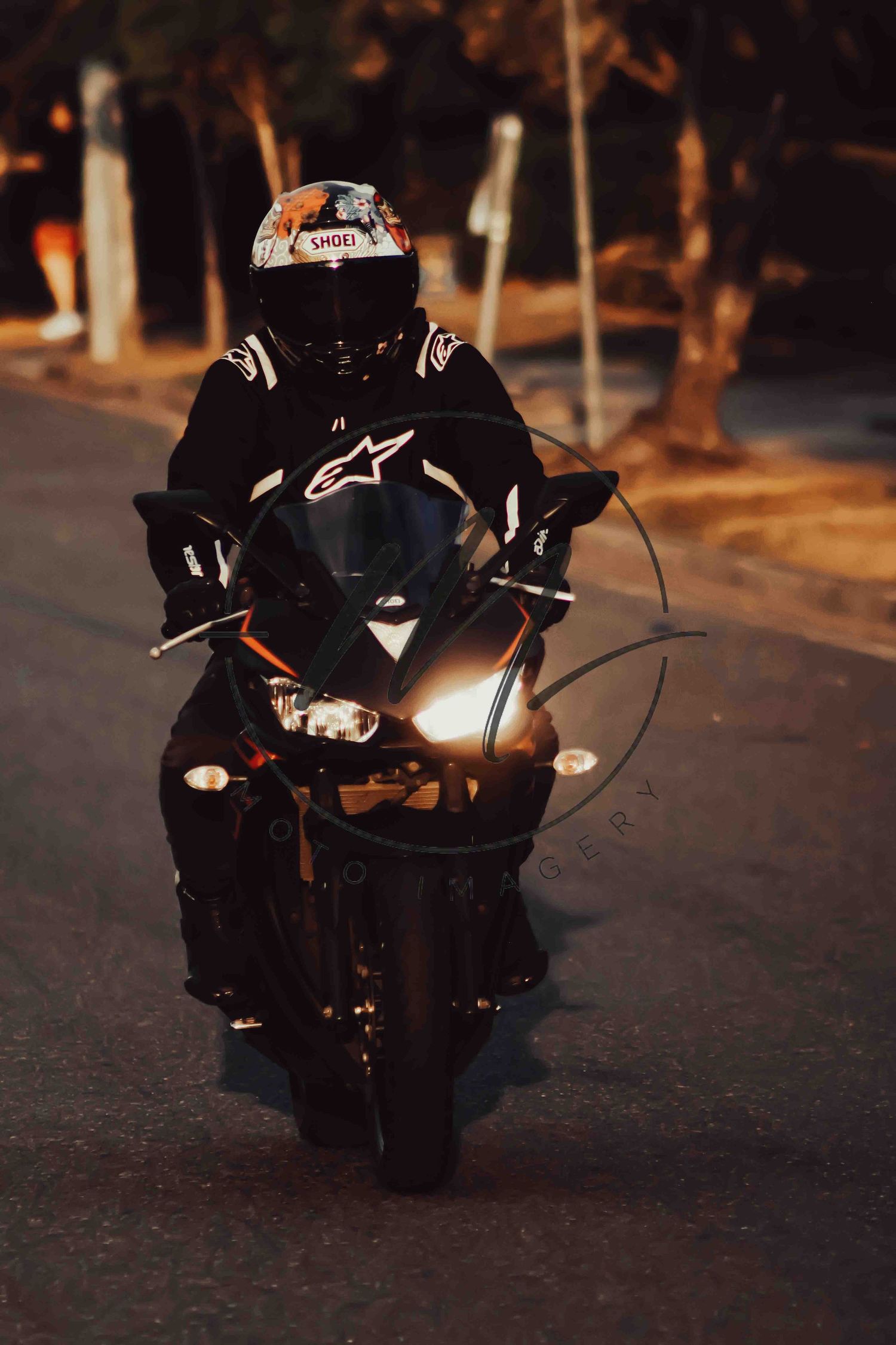 Motorcyclist in black Alpinestars gear and white SHOEI helmet riding a sport bike at night with headlight on.