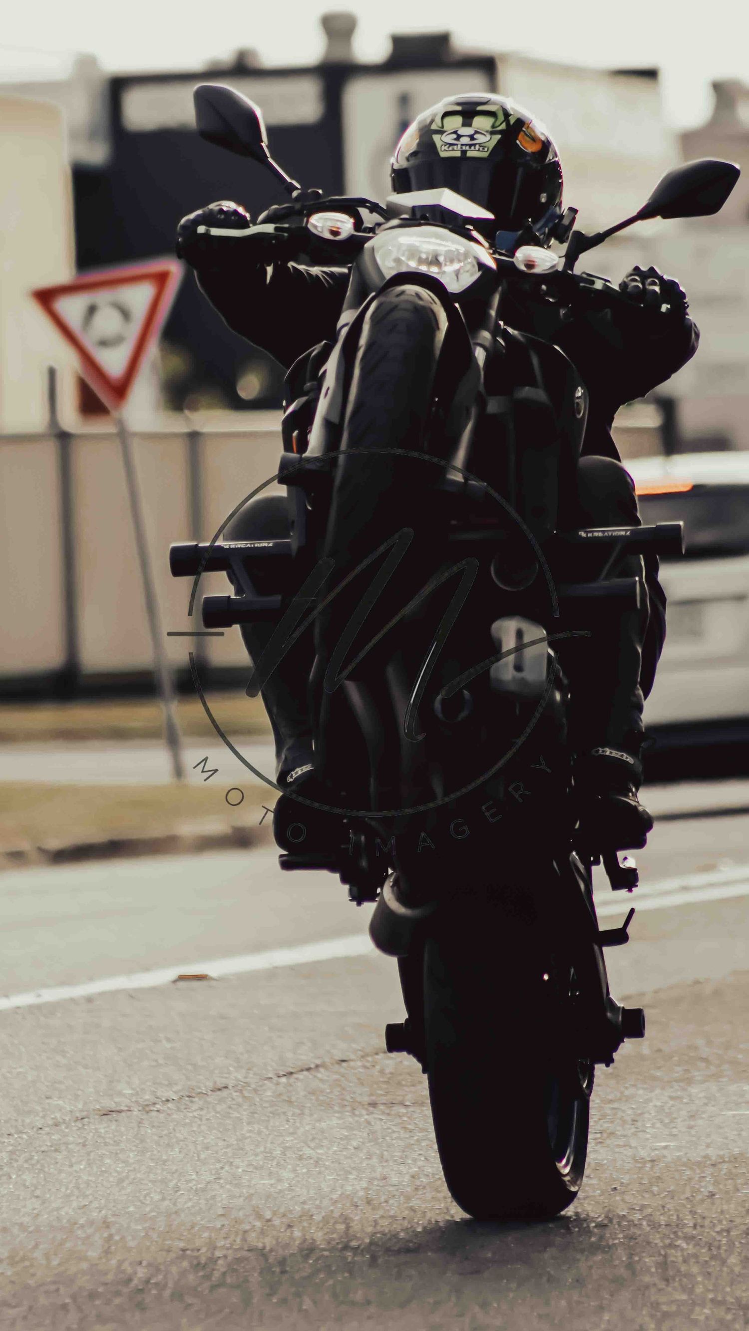 A motorcyclist in black gear and helmet performs a wheelie on a dark sport bike on a city road.