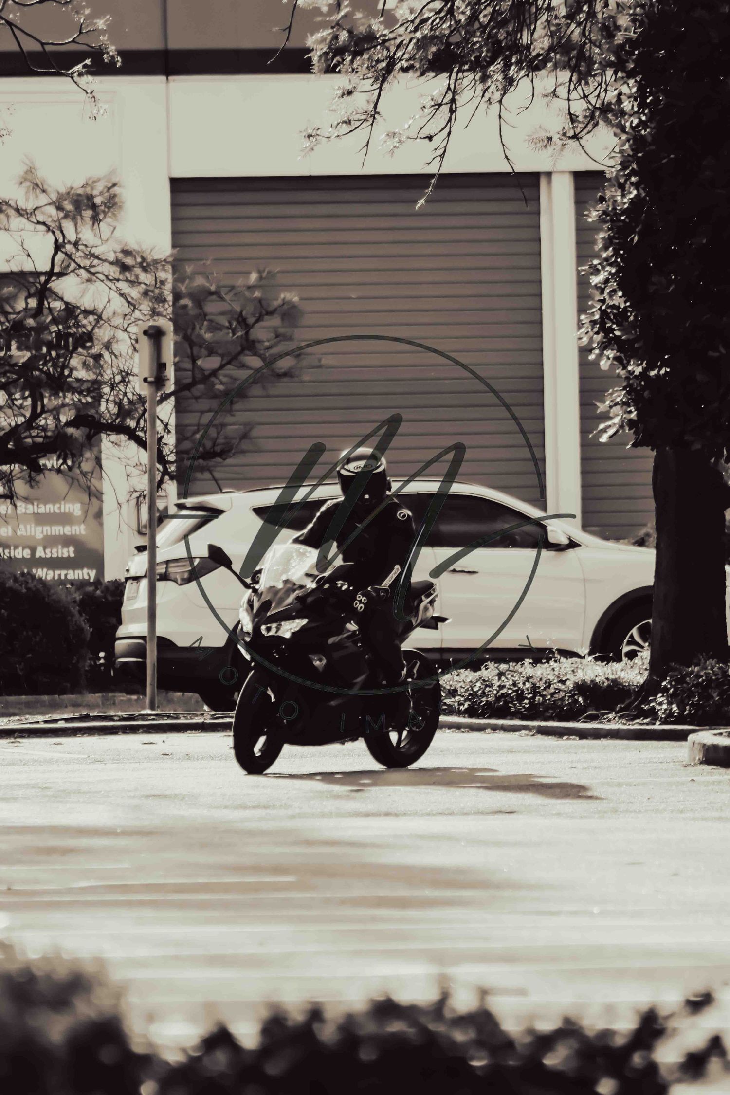 Black and white photo of a motorcyclist riding on a street near a building with a parked car in the background.